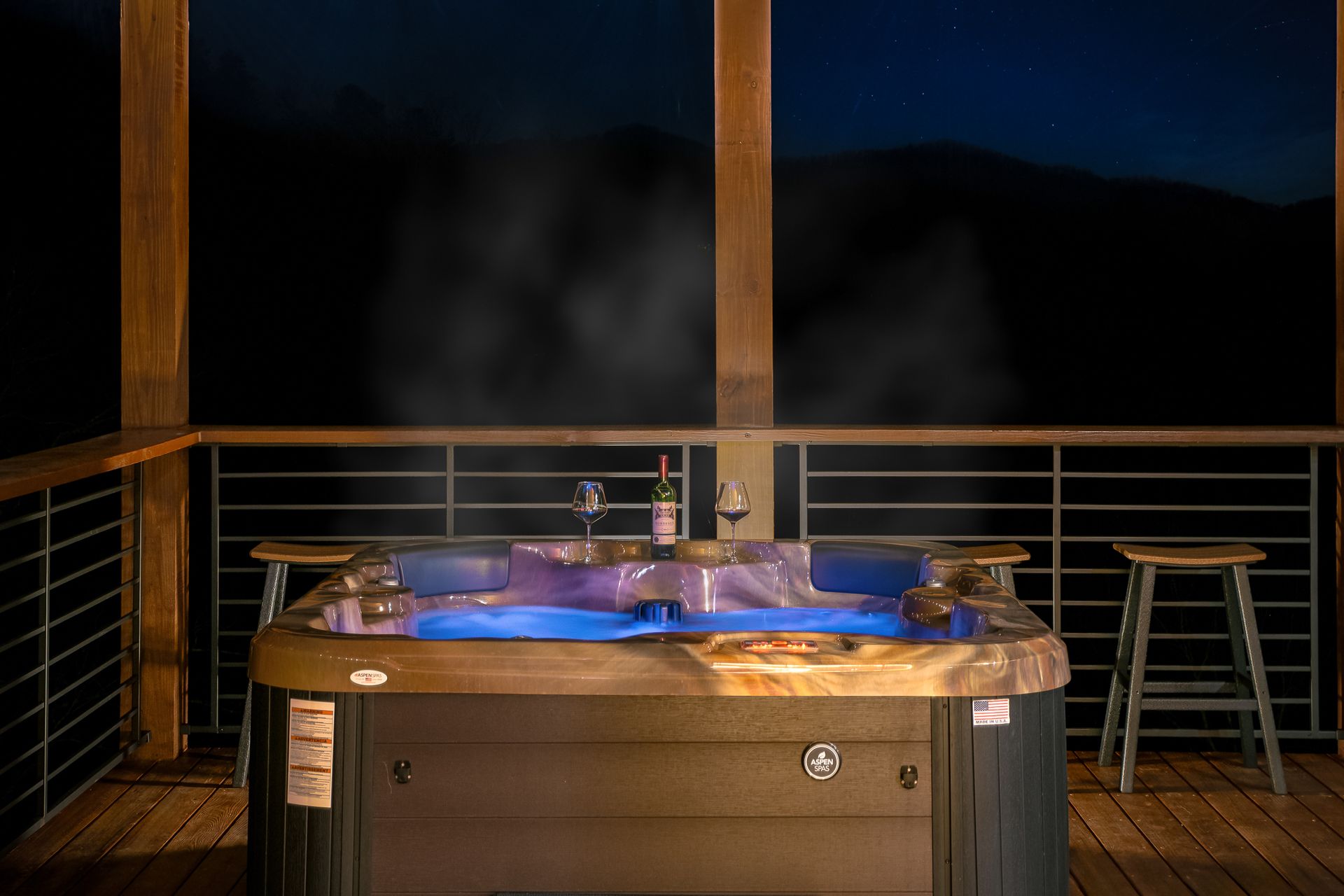 A steaming hot tub sits on a wooden deck at night, with a wine bottle and two glasses placed on the rim.
