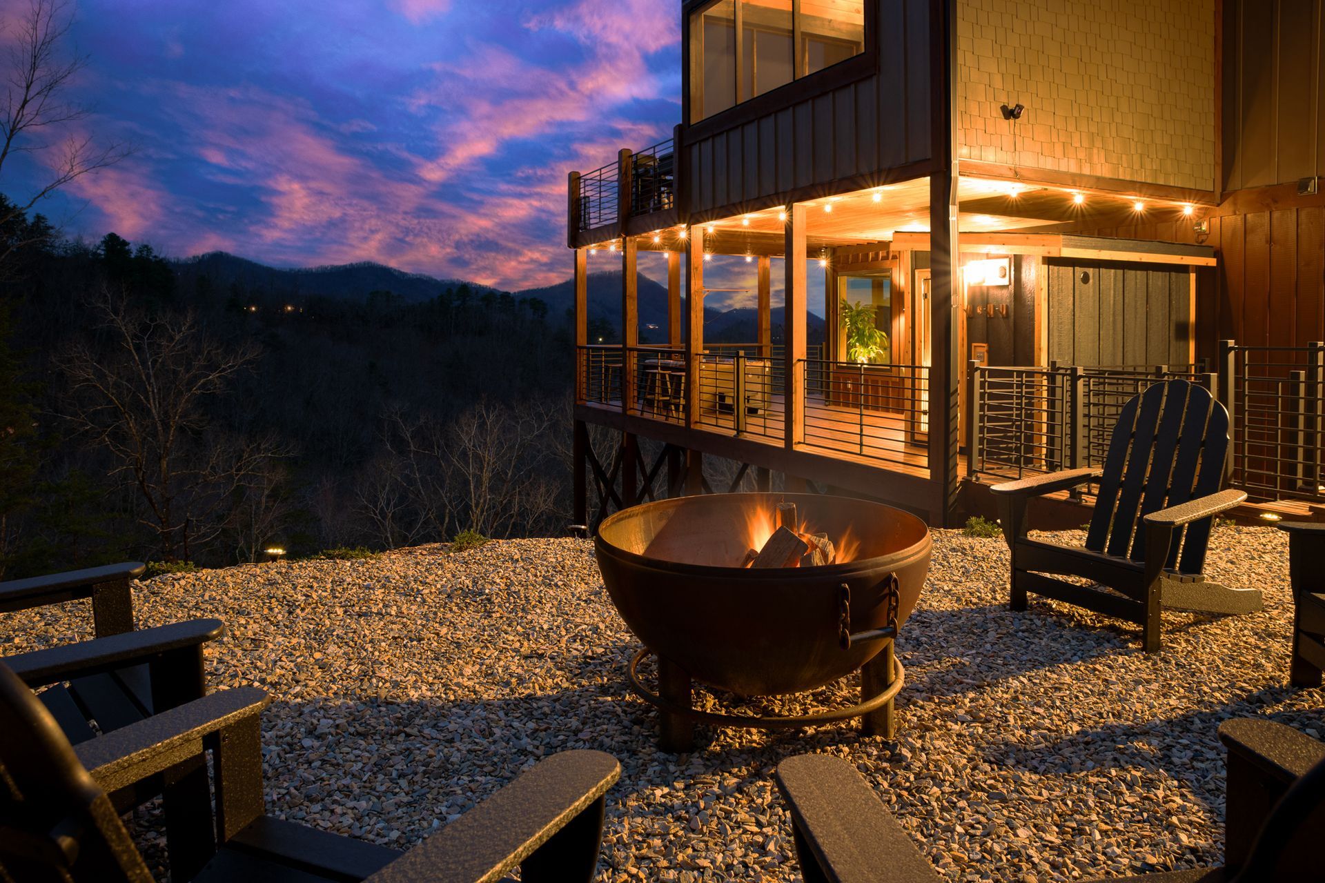 A sunset view of a fire pit surrounded by chairs on a gravel patio beside a lit deck, with mountains in the background.