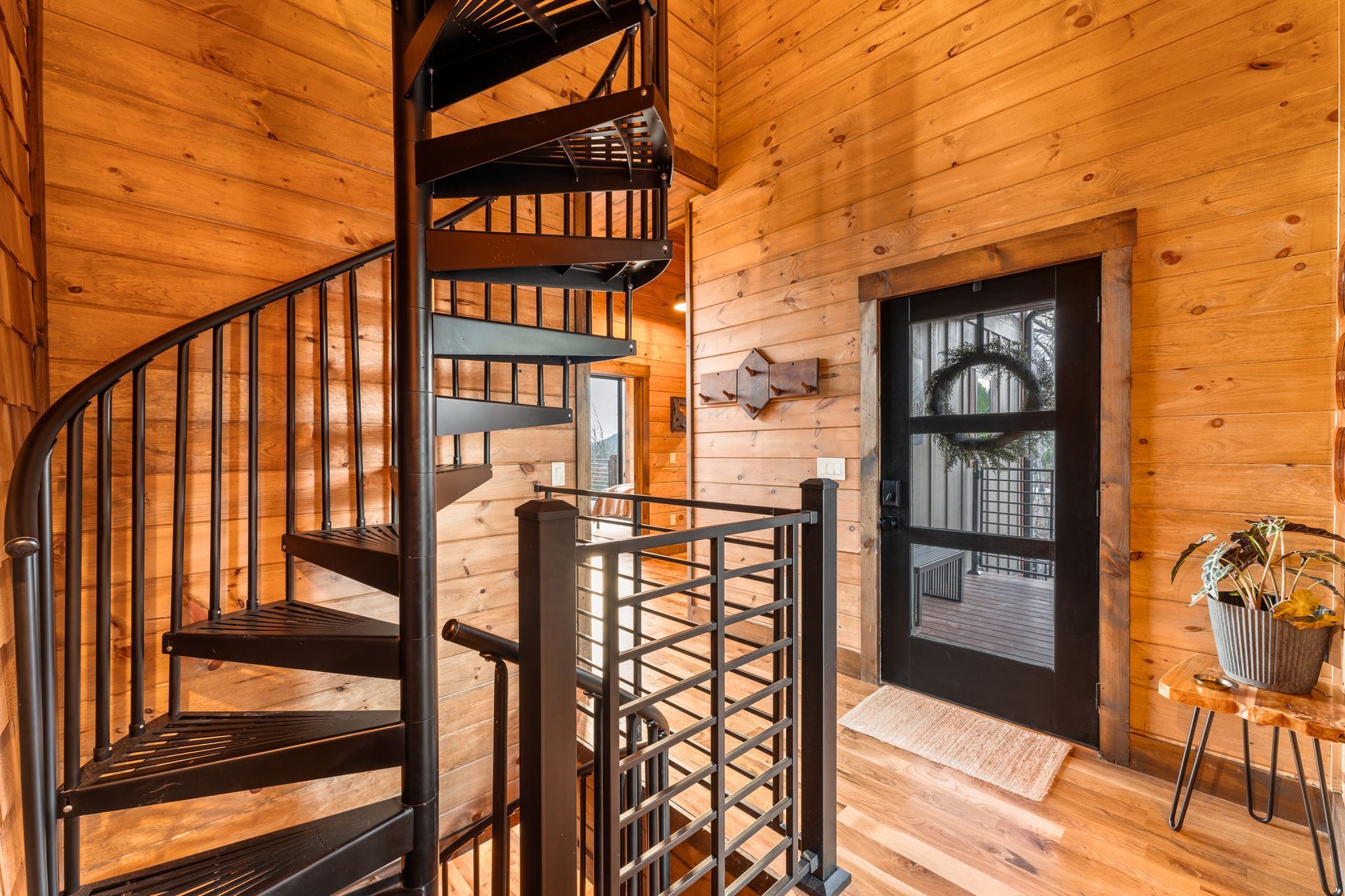 A spiral staircase with black metal railings in a rustic wooden cabin next to a black door with a wreath.