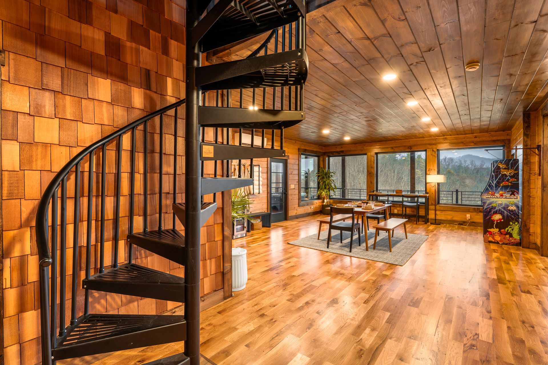 A black spiral staircase in a rustic wood-paneled room with a dining area, wooden floors, and large windows.