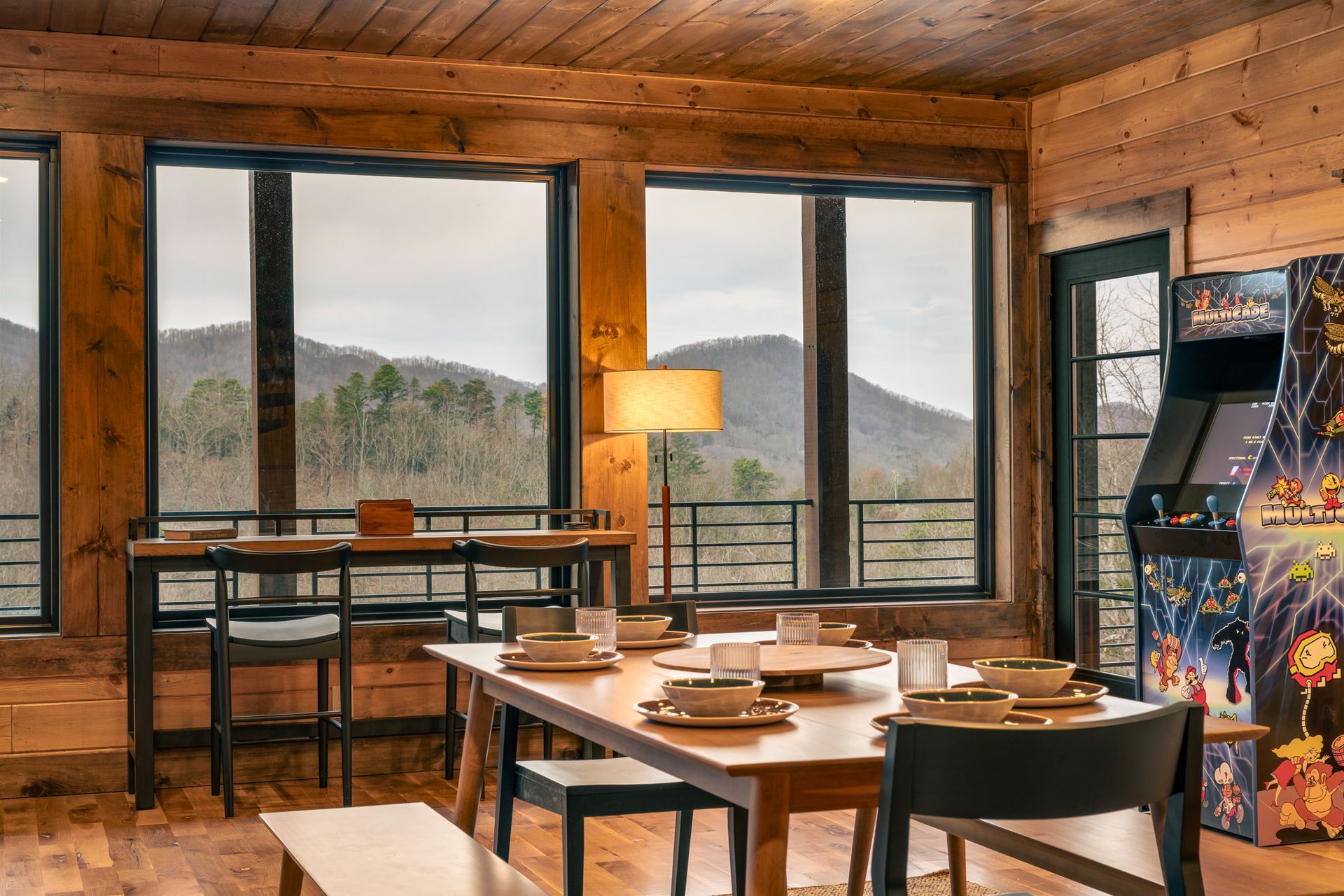 A dining table with chairs and an arcade game in a rustic log cabin with floor-to-ceiling windows overlooking mountains.