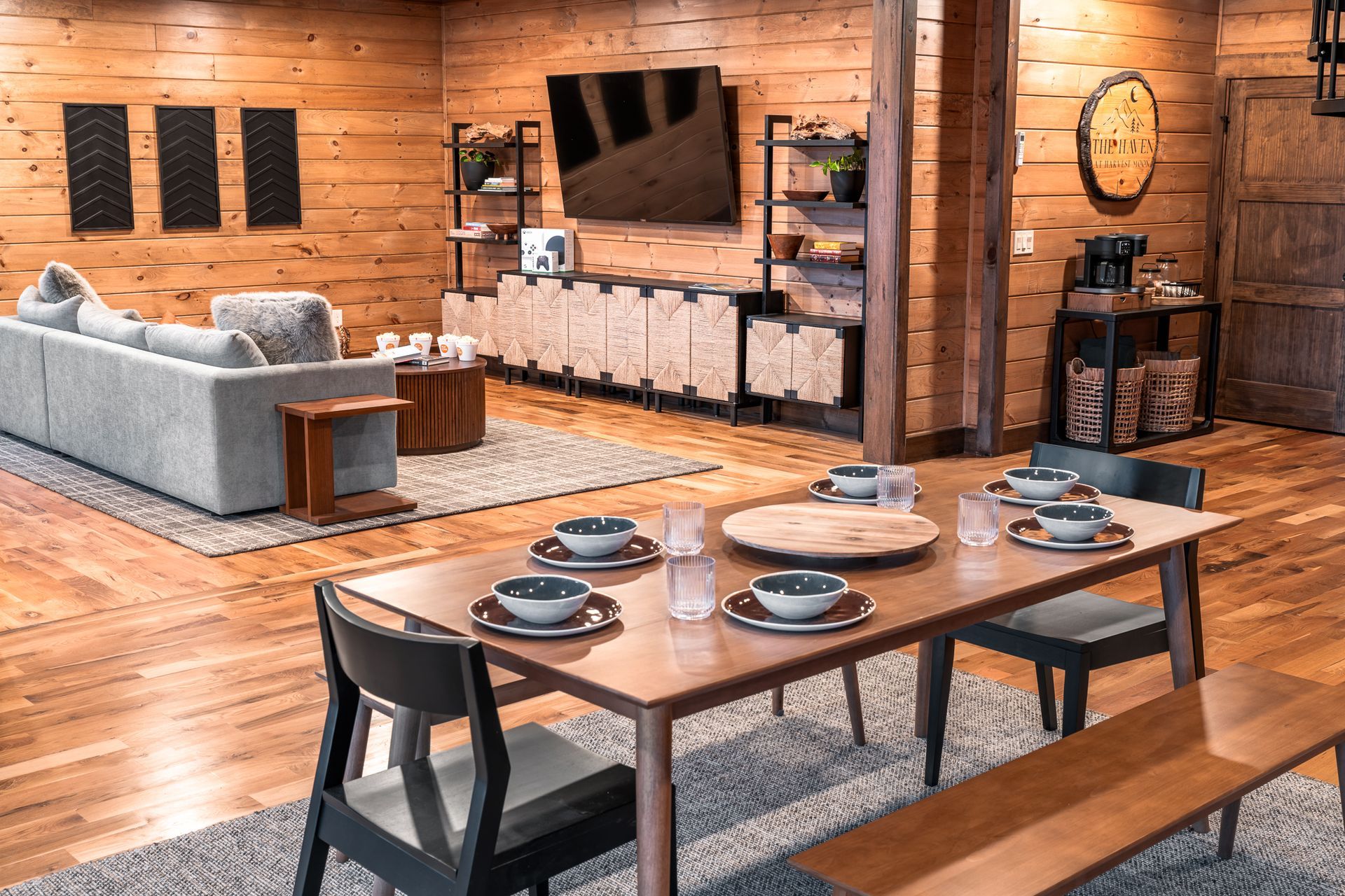 A rustic living and dining area with wood-paneled walls, a gray sofa, and a wooden dining table set for a meal.