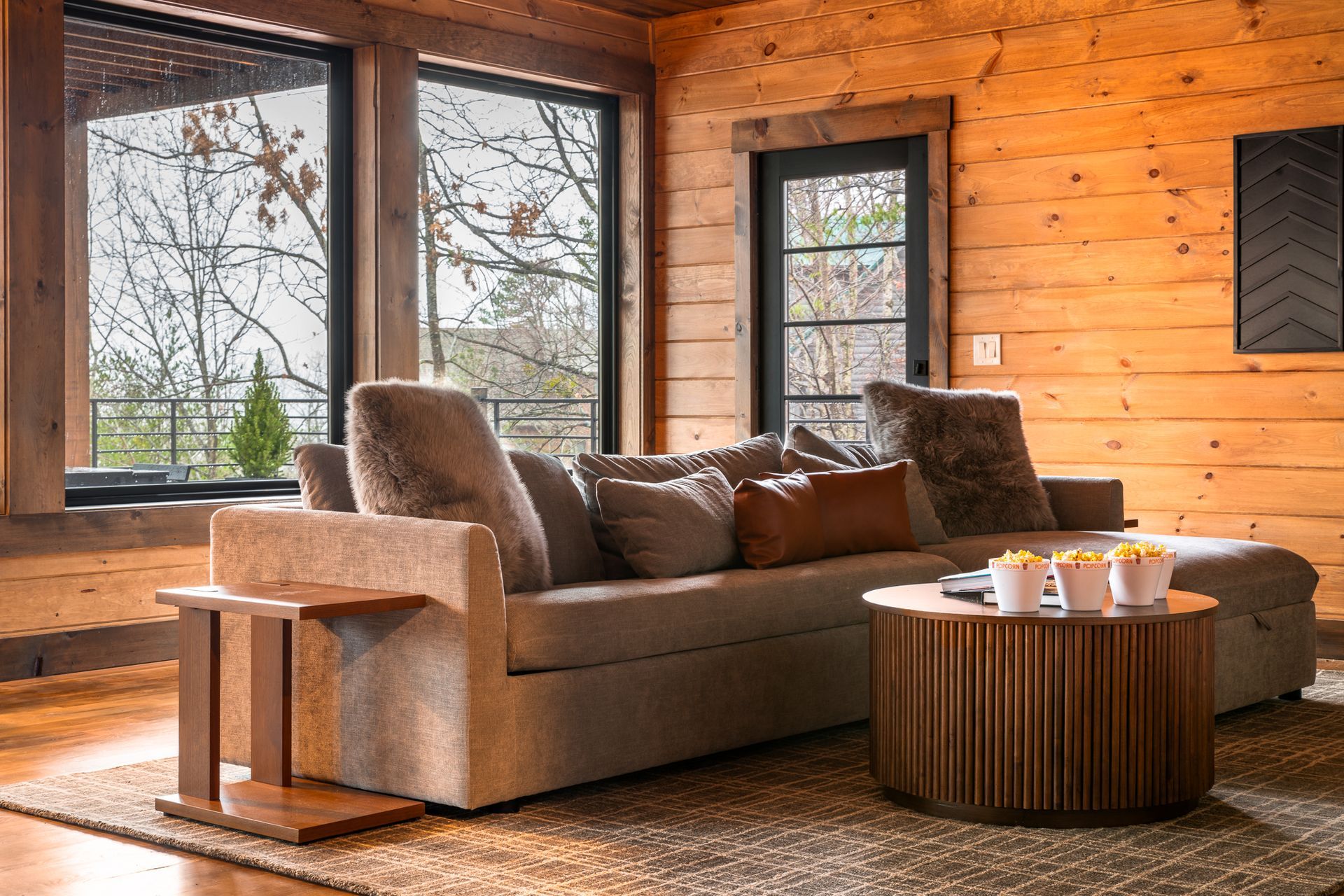 A modern couch with throw pillows and a round slatted coffee table with bowls of popcorn in a wood-paneled living room.