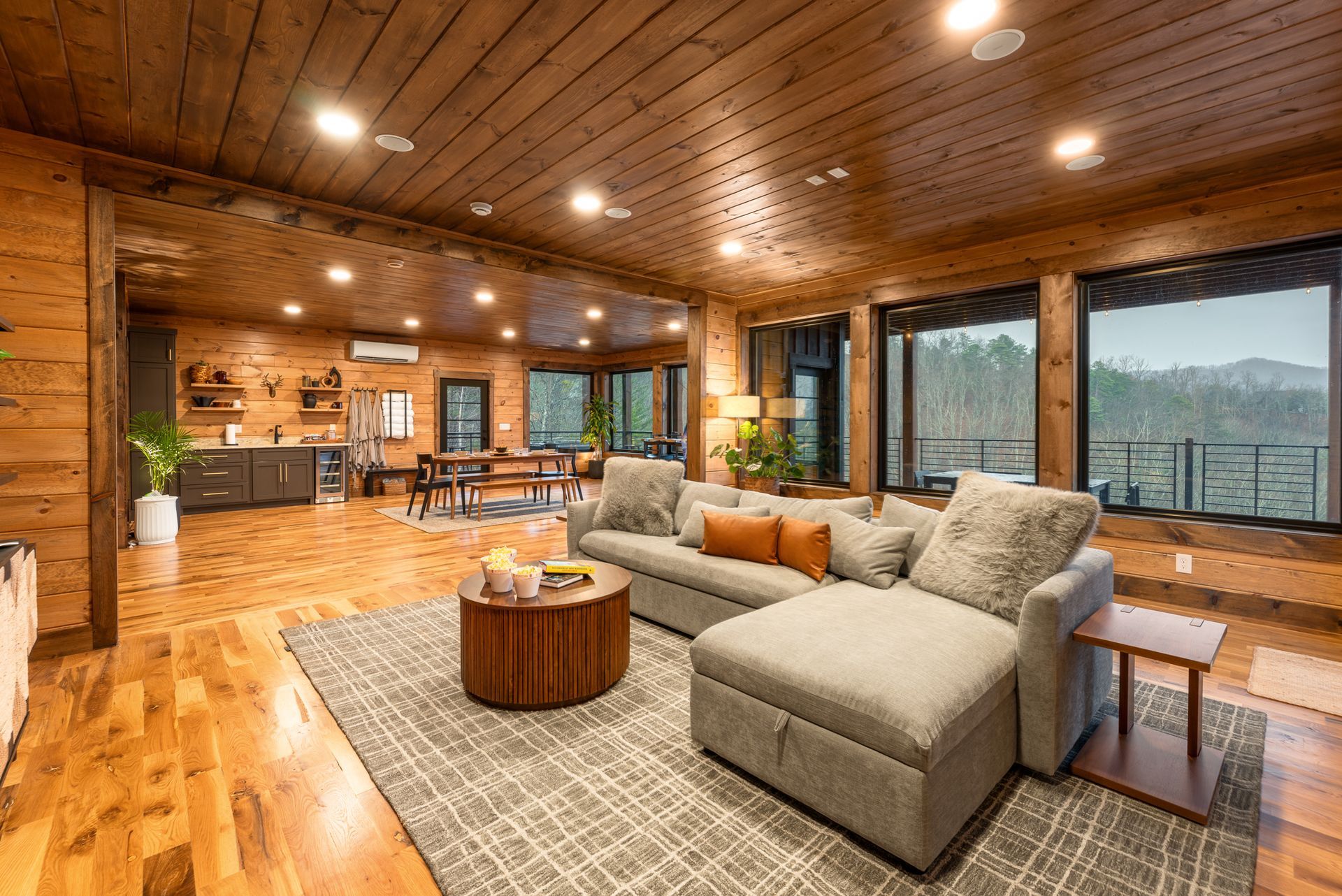 A spacious, wood-paneled cabin living room with a large gray sectional, a circular coffee table, and mountain views.