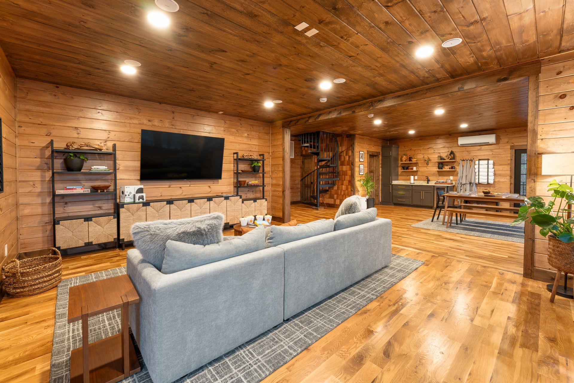 Rustic living room with wood paneled walls, a large grey sofa on a patterned rug, flat-screen TV, and dining area.