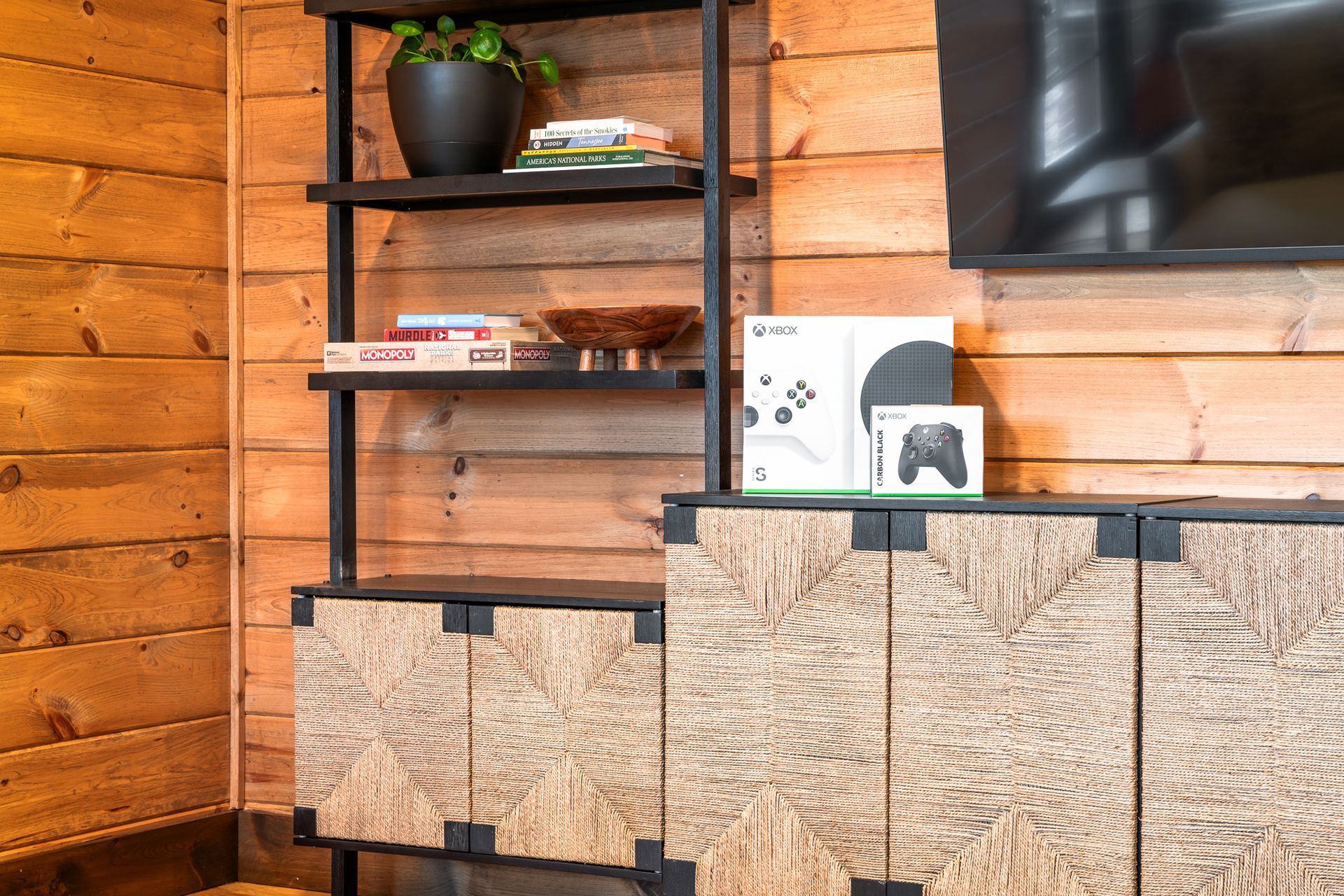 A black metal shelf unit with a houseplant, books, and gaming console boxes next to a wooden wall and media cabinet.