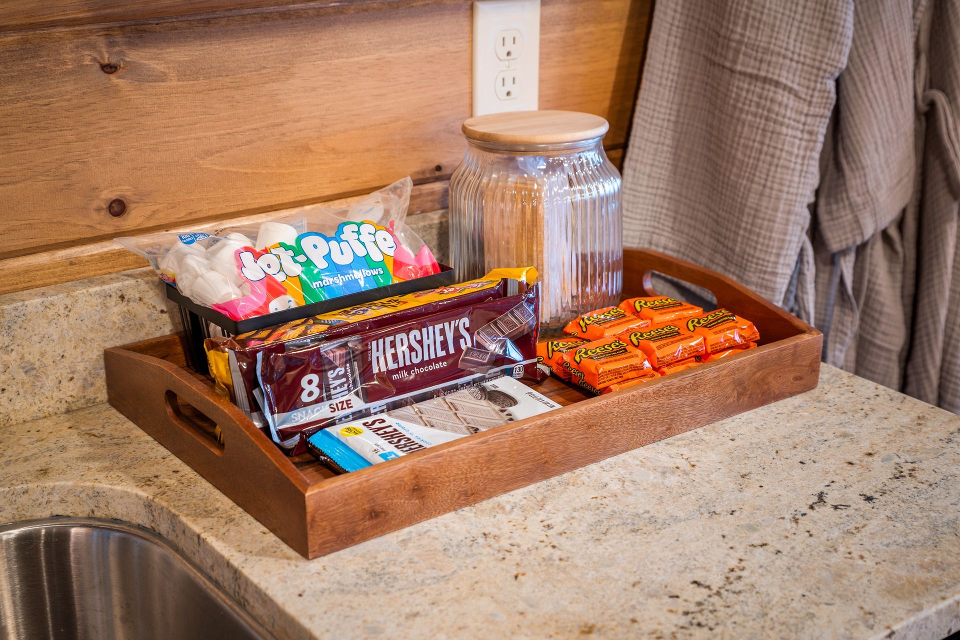 A wooden tray on a kitchen counter holds ingredients for s'mores: Jet-Puffed marshmallows, Hershey's bars, and Reese's.