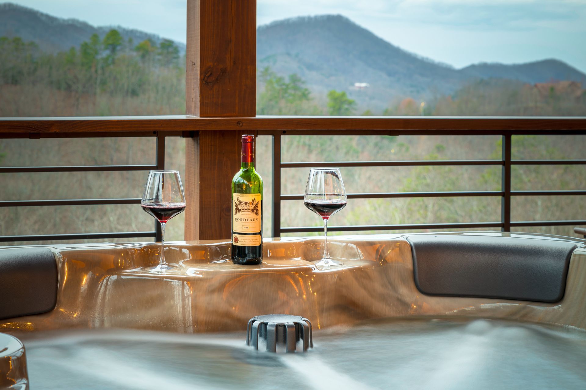 A wine bottle and two glasses of red wine sit on the edge of a steaming hot tub overlooking a scenic mountain landscape.