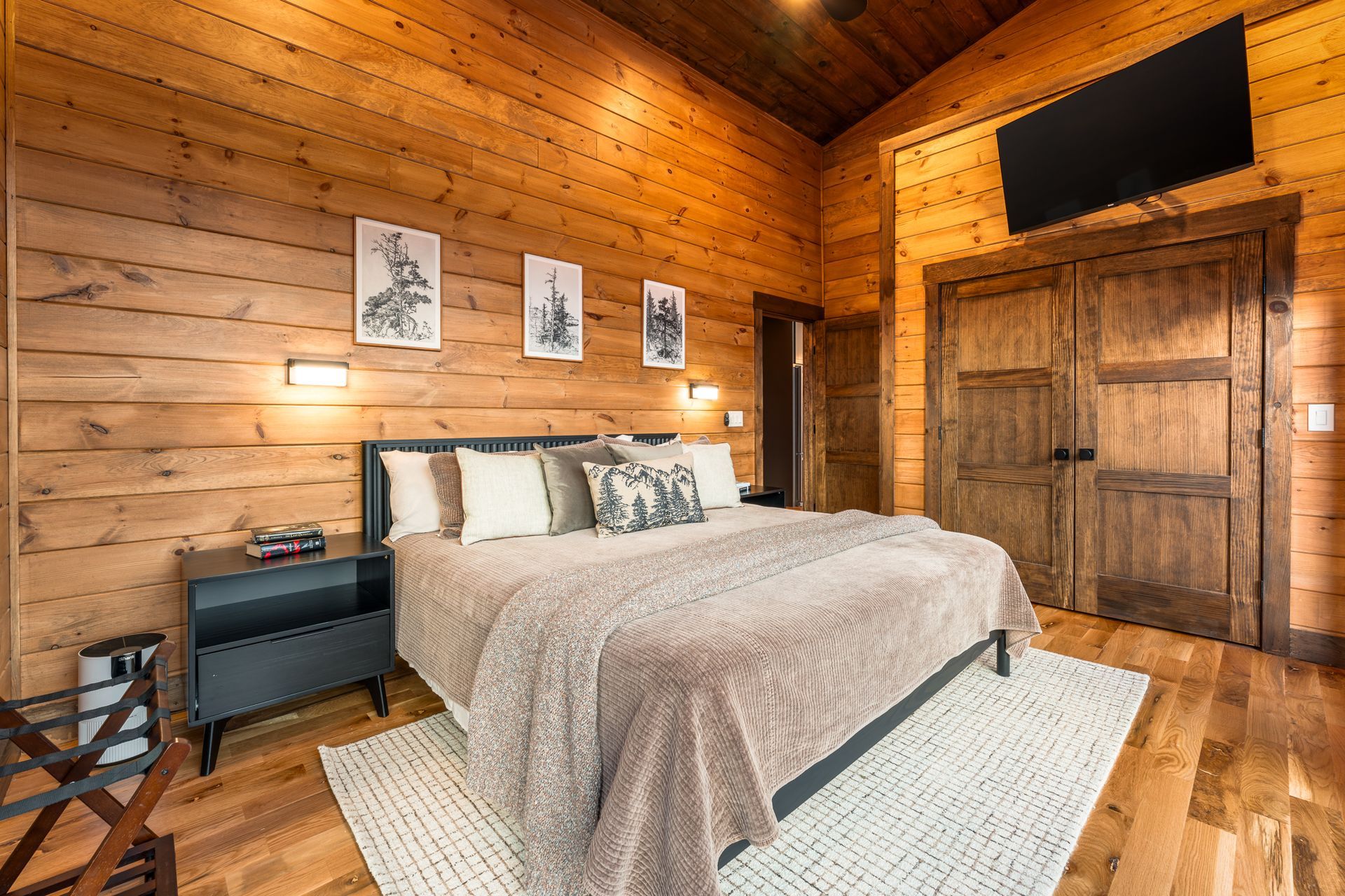 A rustic bedroom with wooden walls, a bed featuring a textured beige blanket and decorative pillows, and a wall-mounted TV.