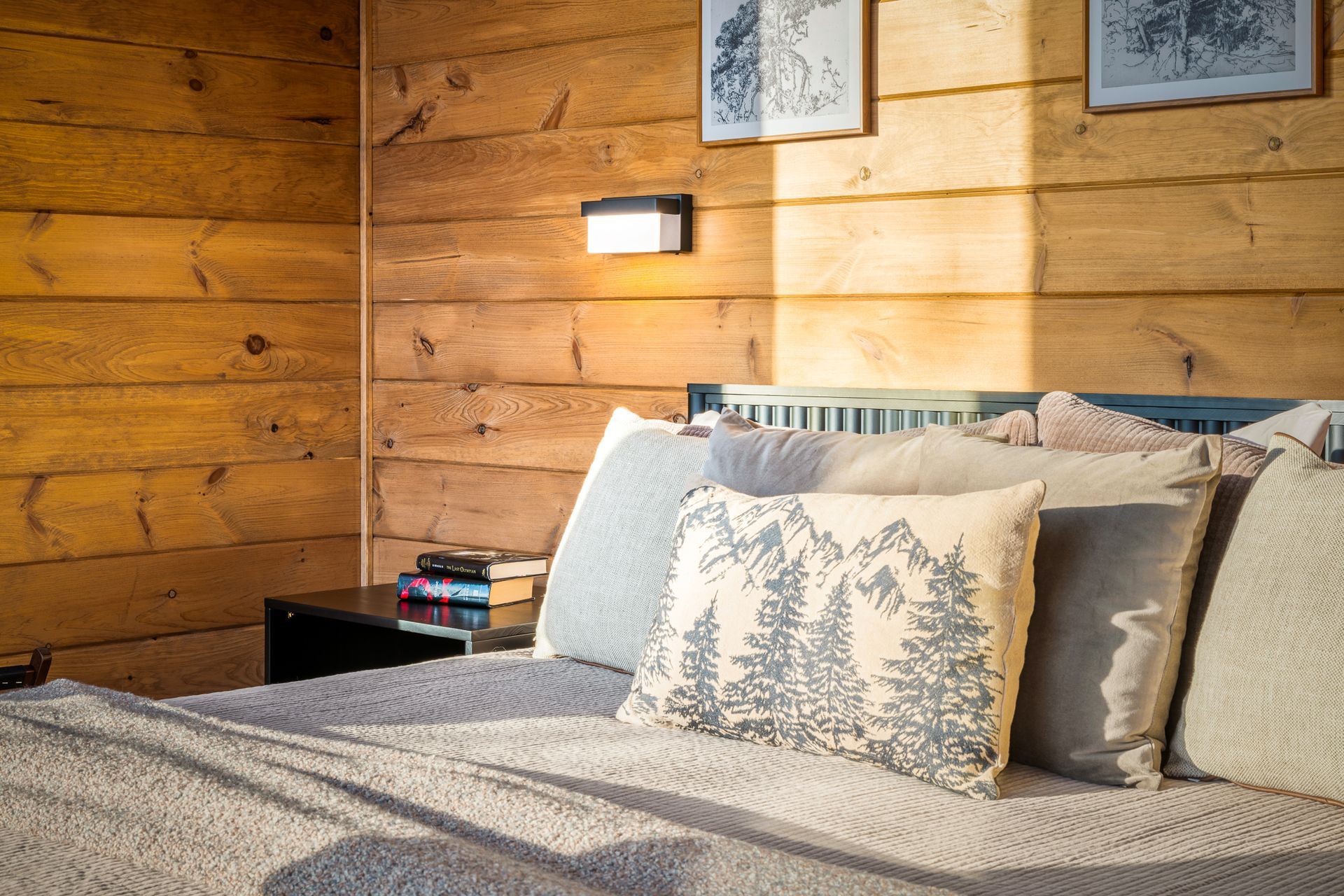 A cozy bedroom with wooden walls, a neatly made bed with patterned cushions, and soft light from a wall-mounted lamp.