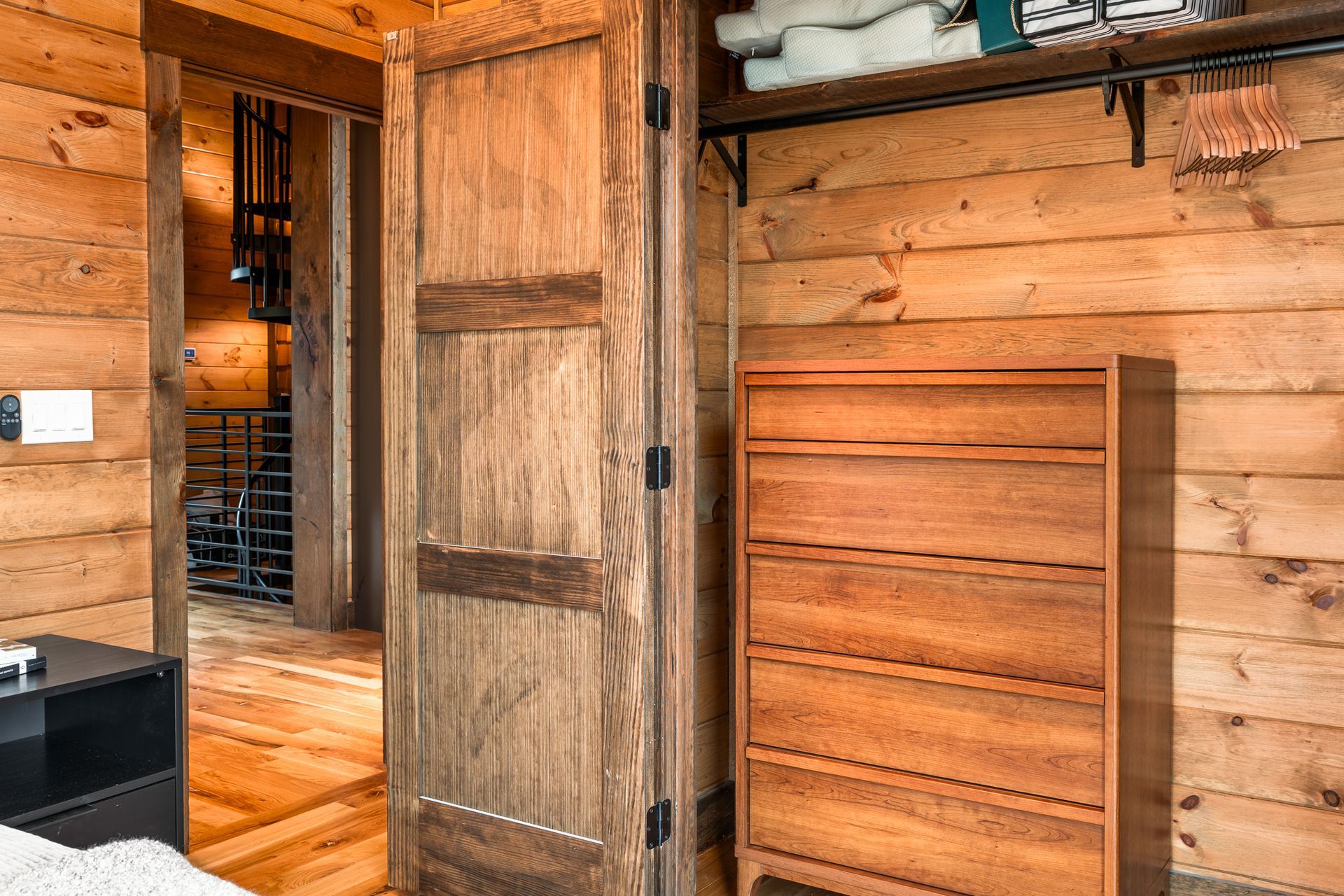 A wooden dresser stands against a log wall next to a wooden door leading to a hallway with a staircase.