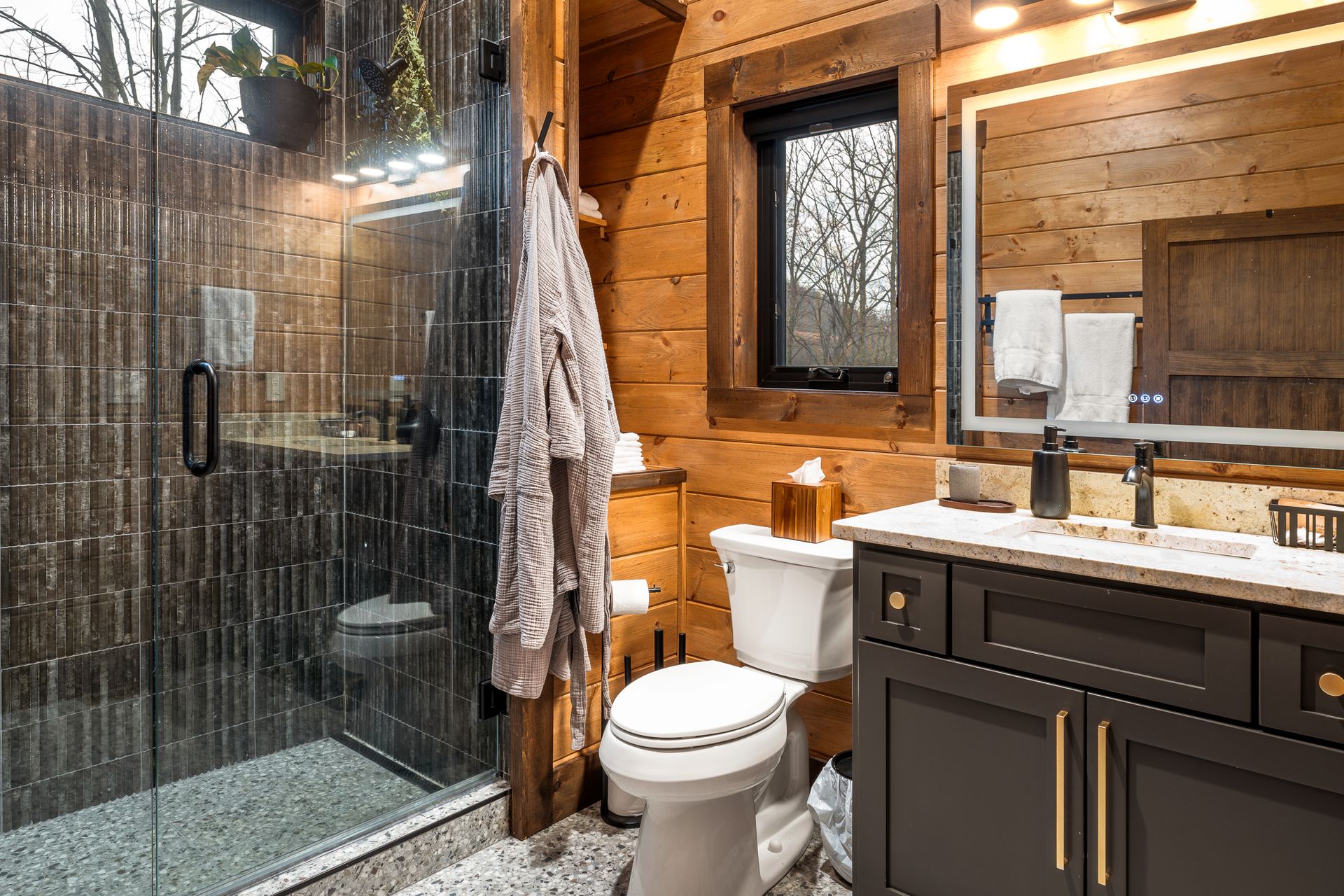 A rustic bathroom with wood walls, a glass shower, a white toilet, and a dark vanity with a modern mirror.