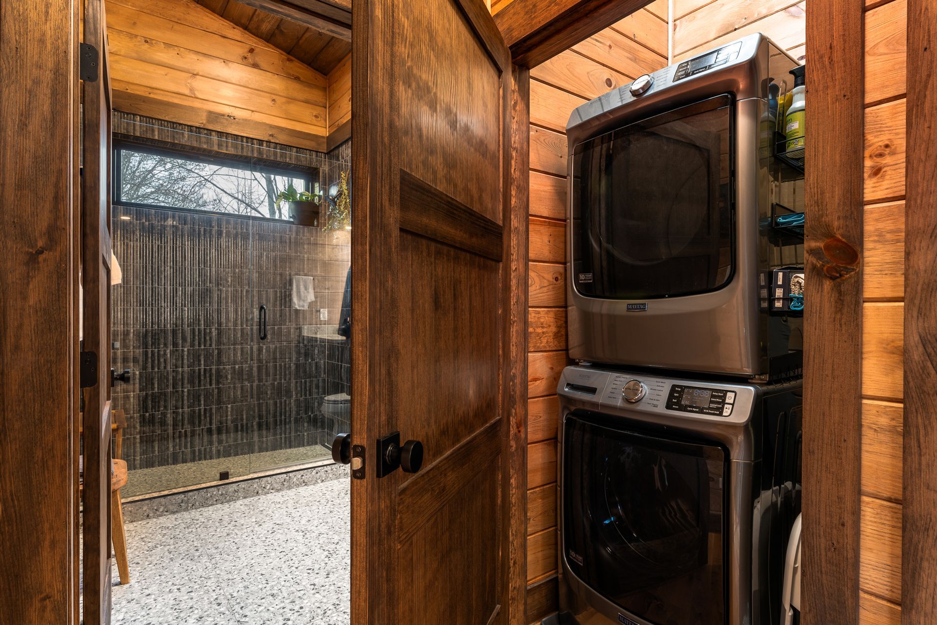 A stacked washer and dryer unit beside a wooden door opening into a bathroom with a glass shower stall.