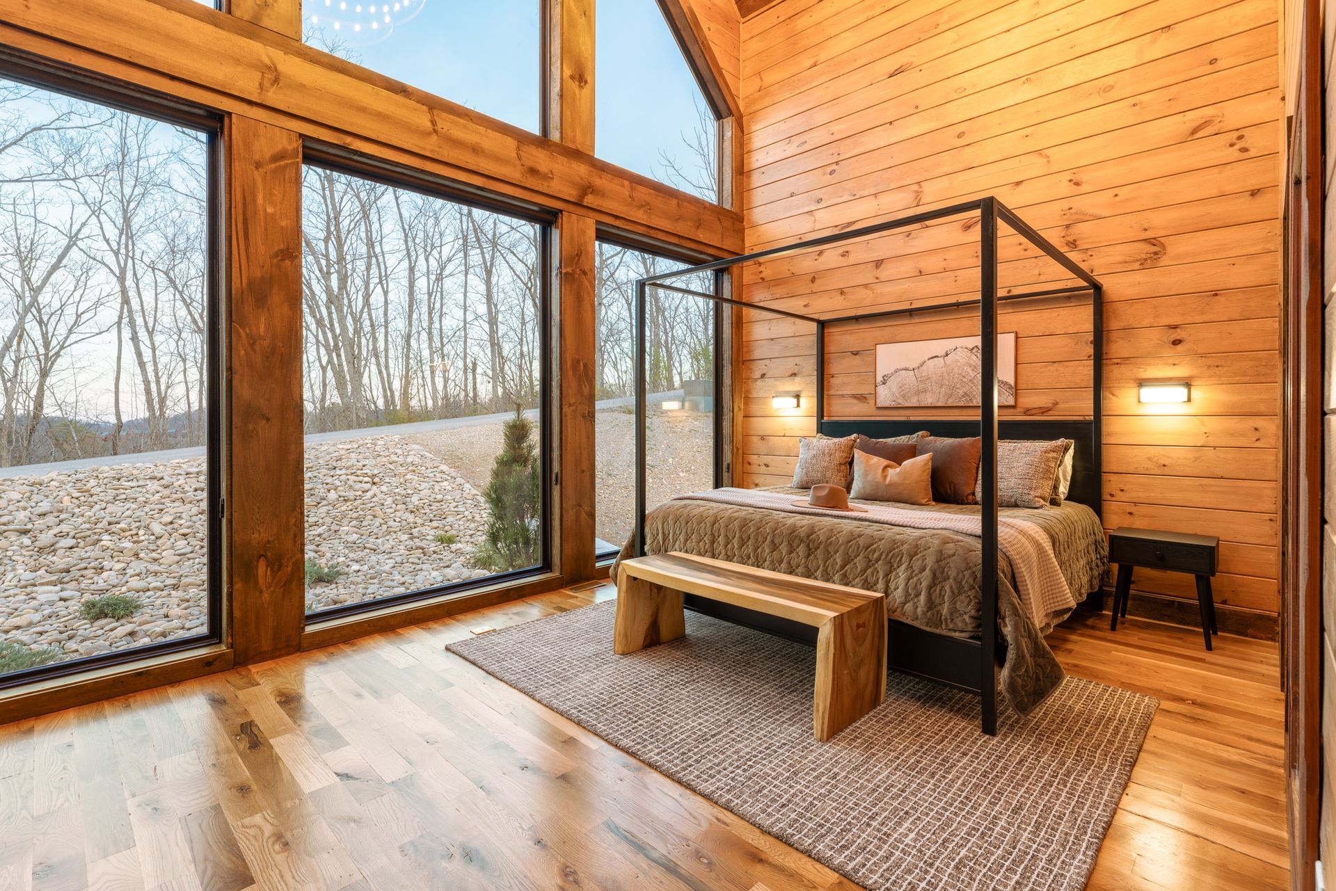 A sunlit wooden bedroom featuring a canopy bed, a rustic wooden bench, and large floor-to-ceiling windows viewing woods.