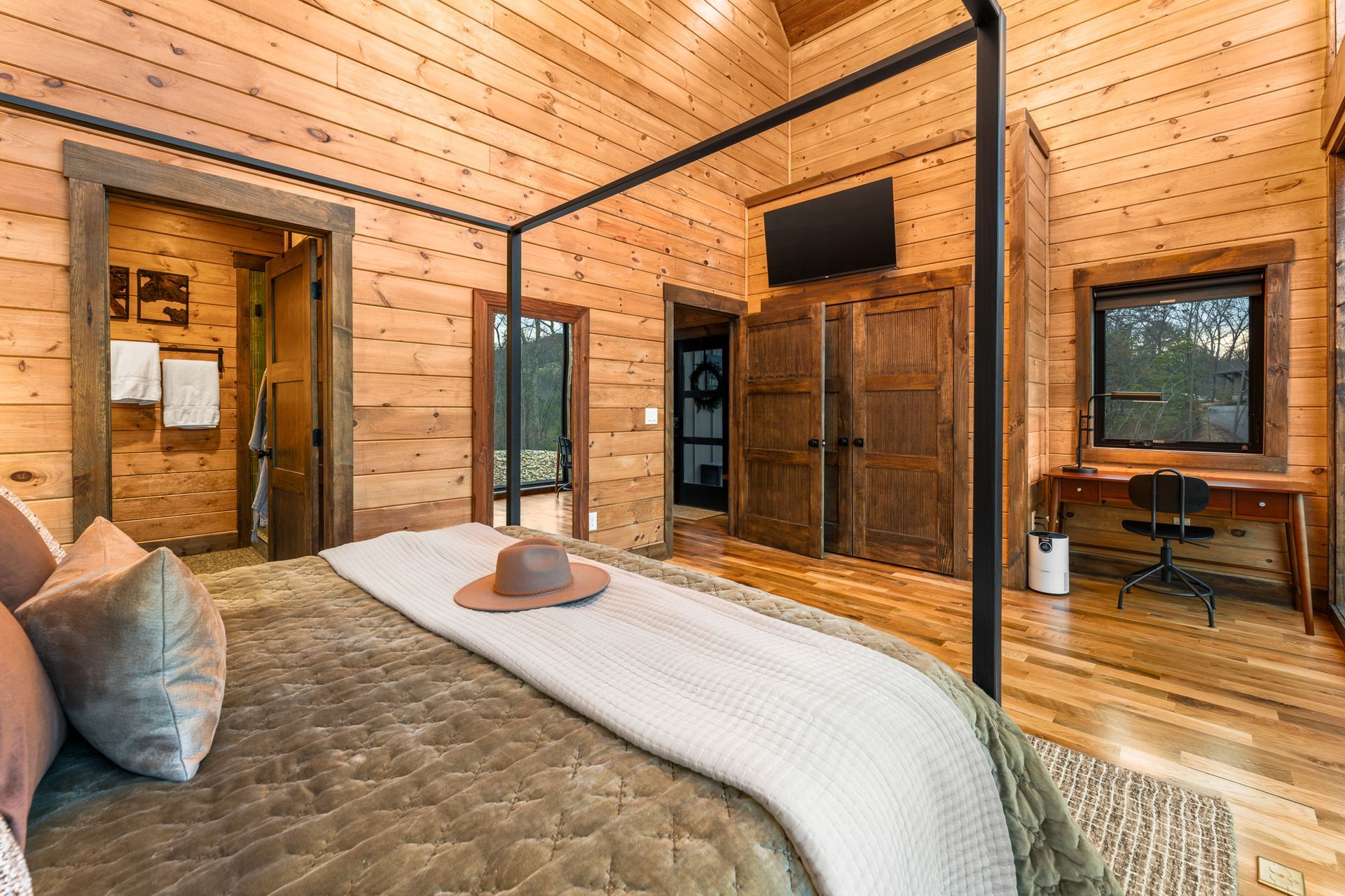 A rustic bedroom with wood paneled walls, a four-poster bed, a desk in the corner, and a television mounted on the wall.