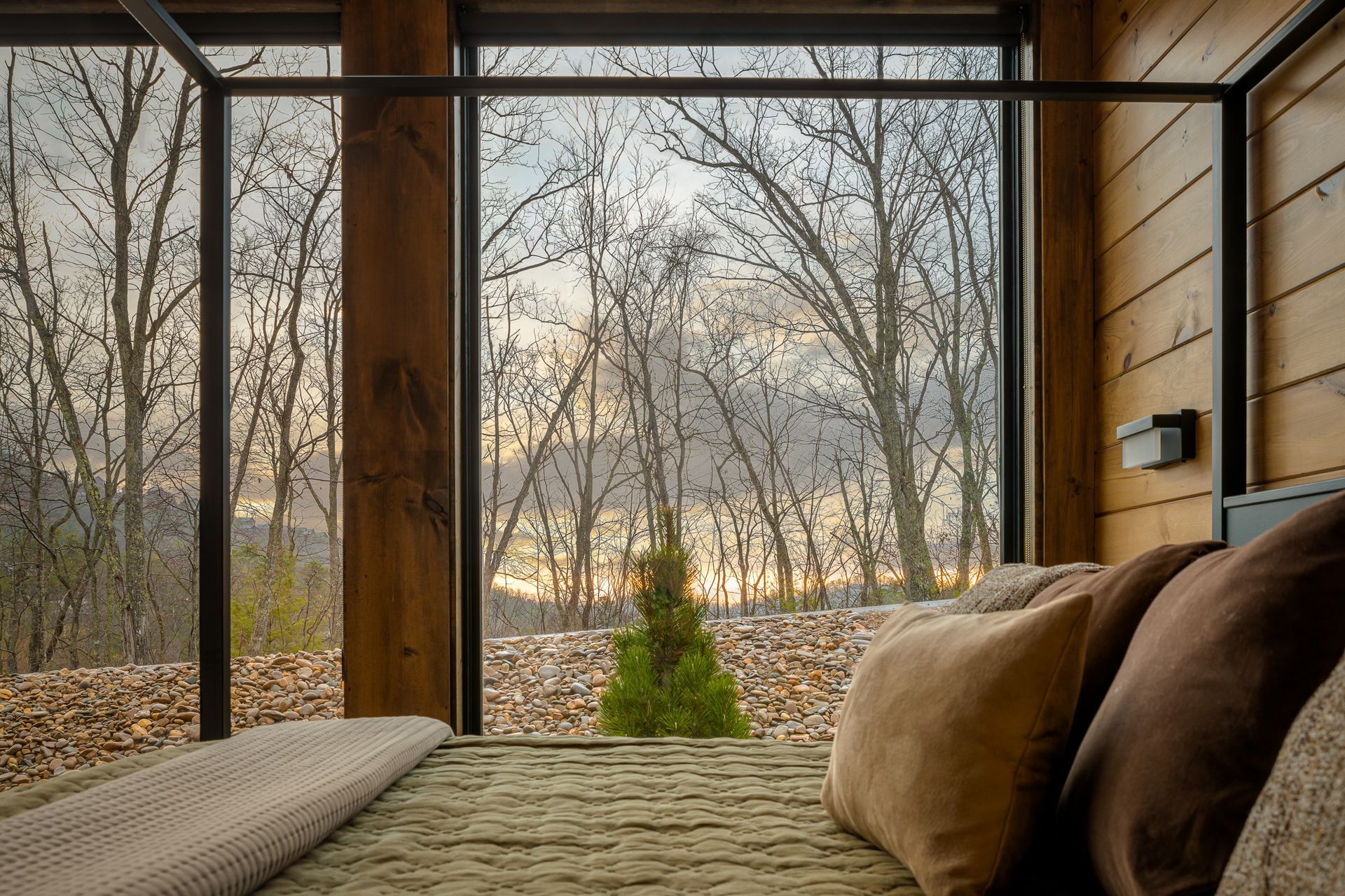 A view from a bed inside a cabin, with large windows looking out over a forest landscape at sunset.