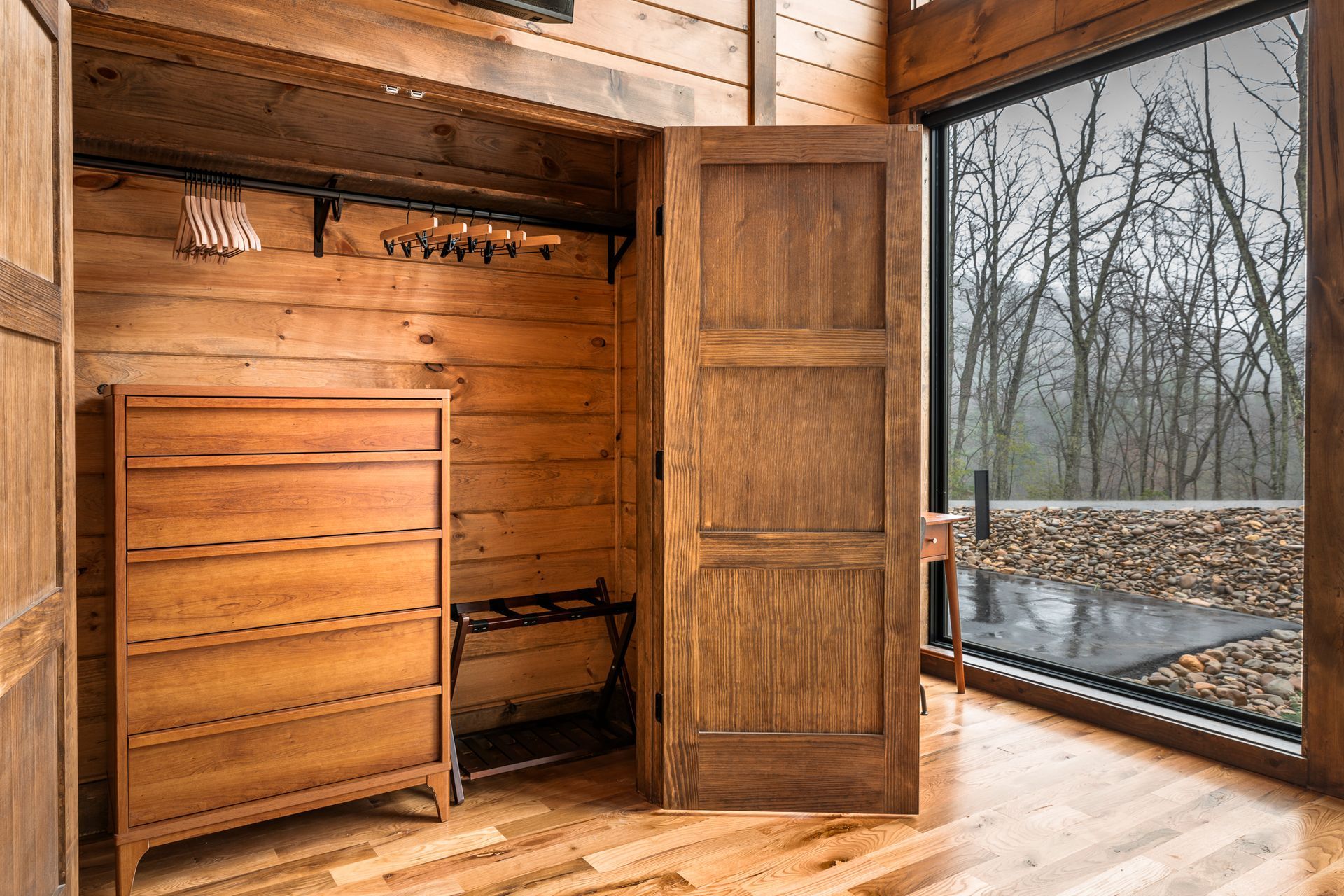 A wooden dresser sits inside an open closet beside a large window looking out at a forest.