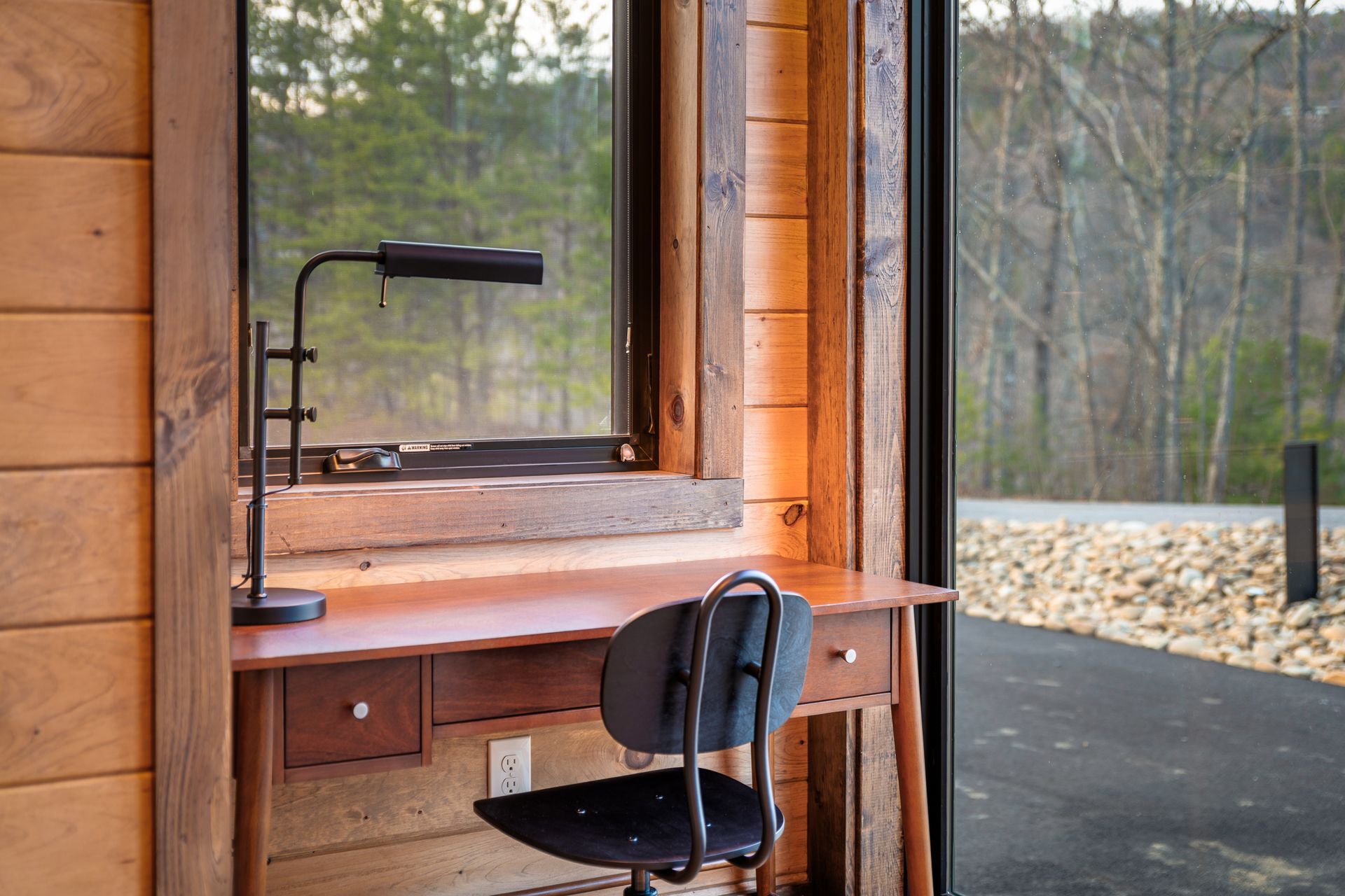 A wooden desk with a black lamp and chair sits next to a window overlooking a tree-lined driveway in a rustic room.