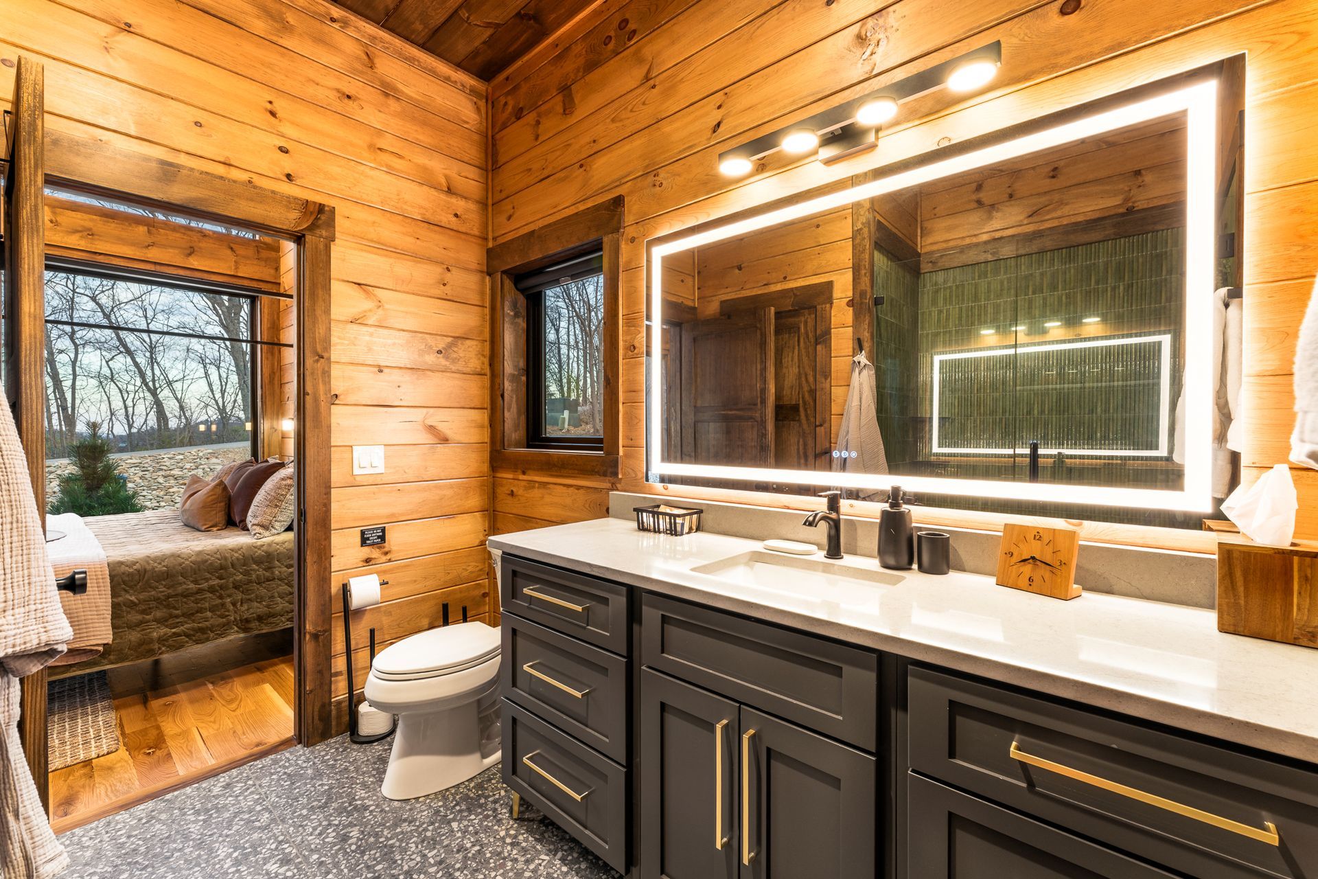 A rustic bathroom with log walls, a dark grey vanity, a large lighted mirror, and a view into a cozy bedroom.