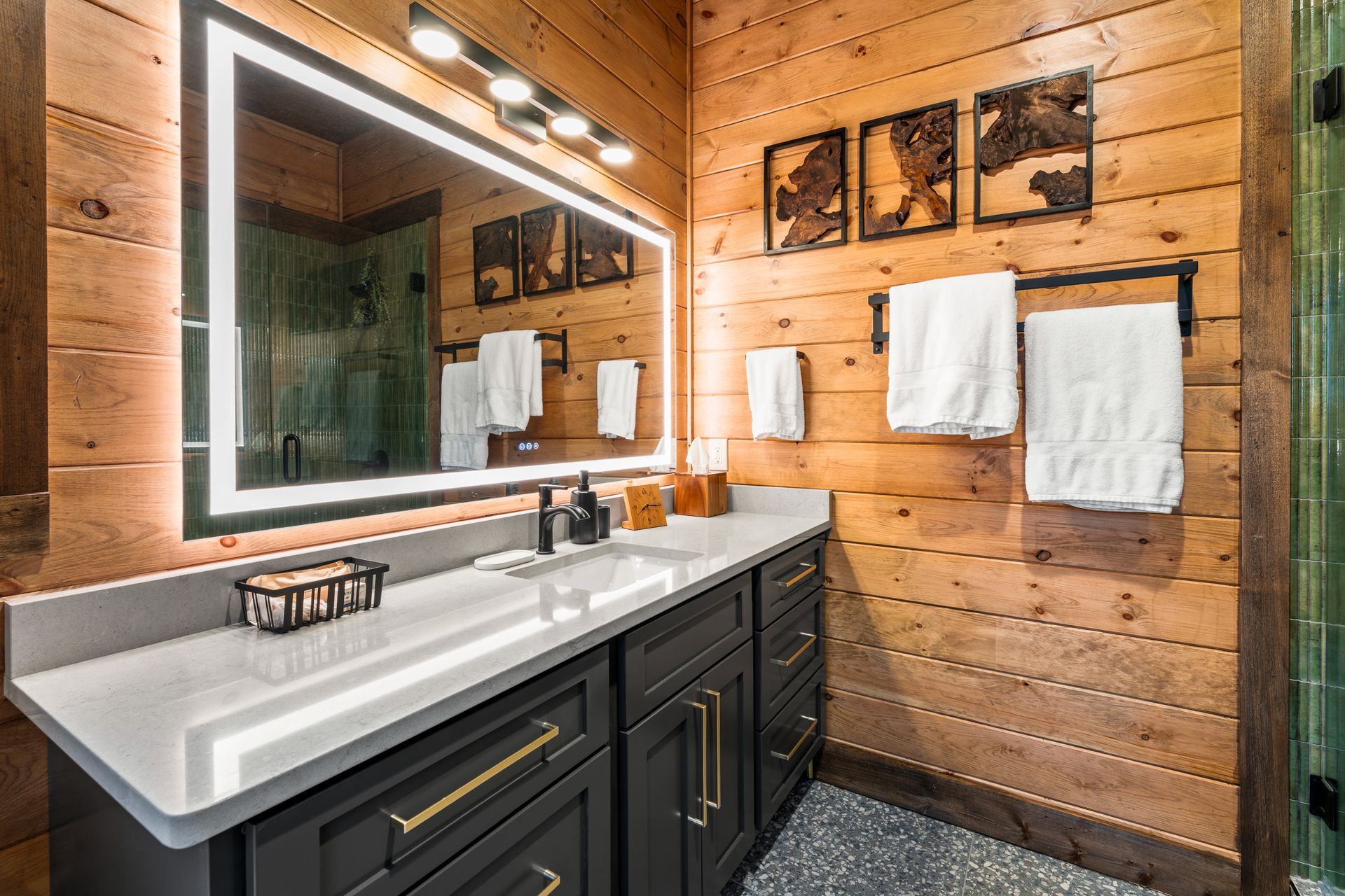 A modern vanity with a lit mirror, dark cabinetry, and white towels on a towel rack against wood-paneled walls.