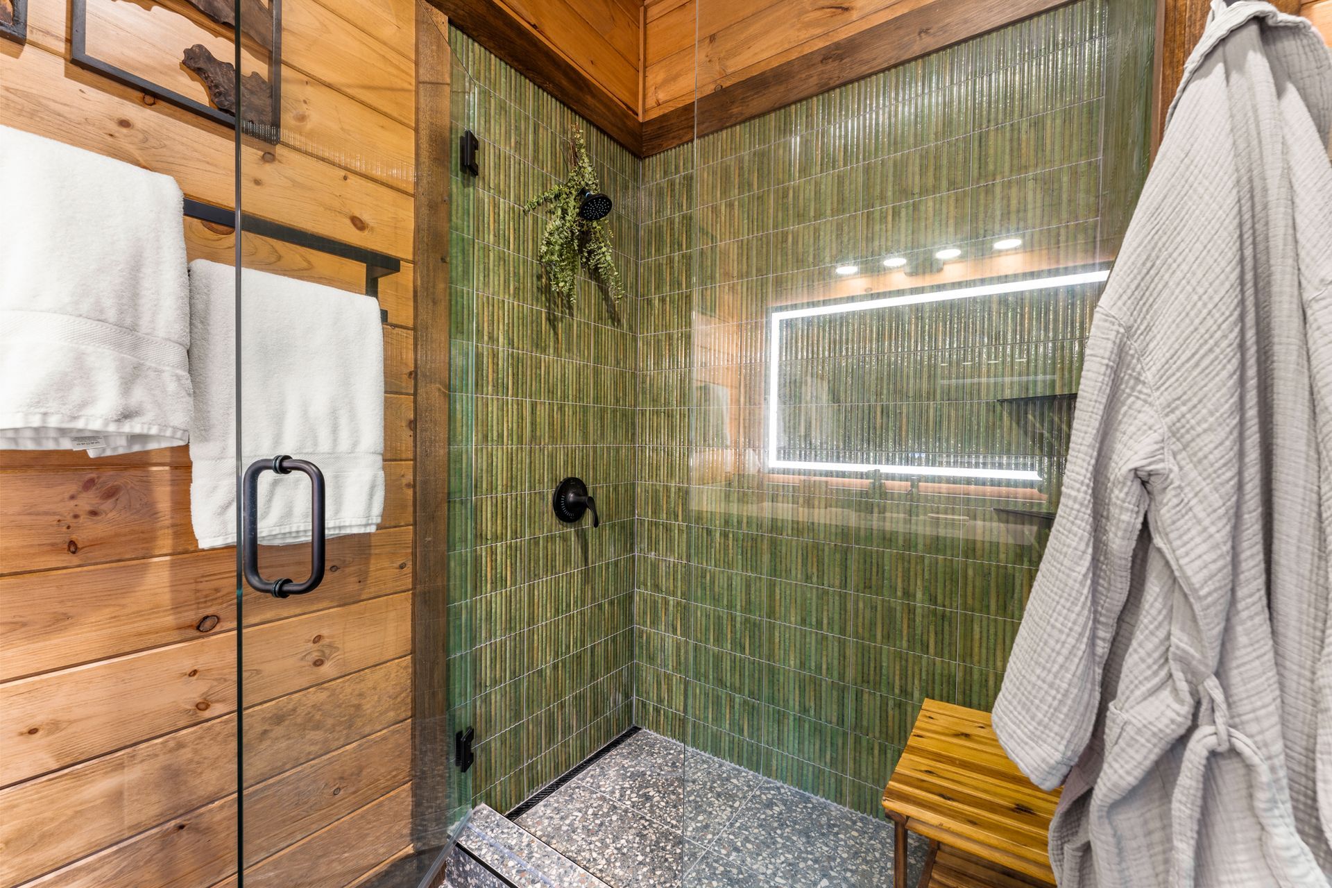 A bathroom shower with sage green vertical tiles, a glass door, wooden walls, a light-up mirror, and a wood bench.