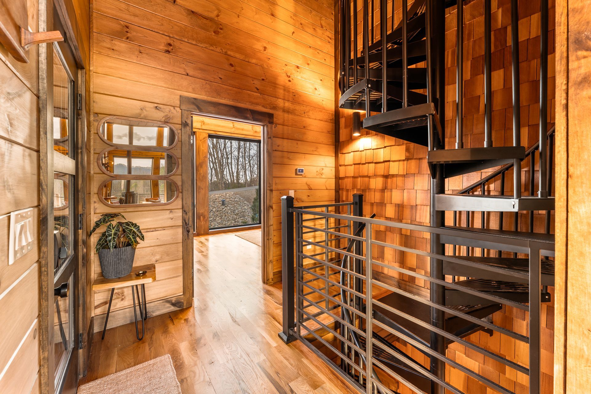 A hallway with wood walls and floors, featuring a black metal spiral staircase, a small plant, and a view into a room.