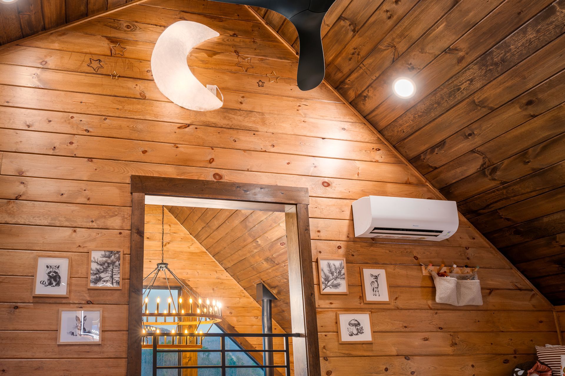 A rustic, wood-paneled loft interior features a crescent moon wall light, framed art prints, and a wall-mounted AC unit.