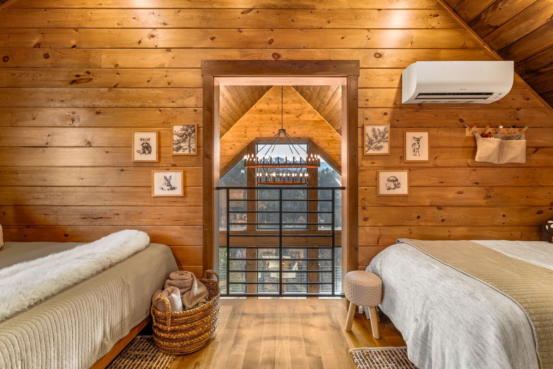 A cozy loft bedroom with wood paneled walls, two beds, a basket of blankets, and a central doorway overlooking a lower level.