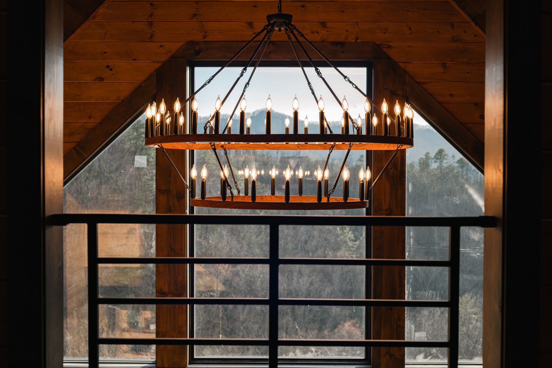 A large, two-tiered rustic chandelier hangs in front of a tall window overlooking a wooded mountain view.