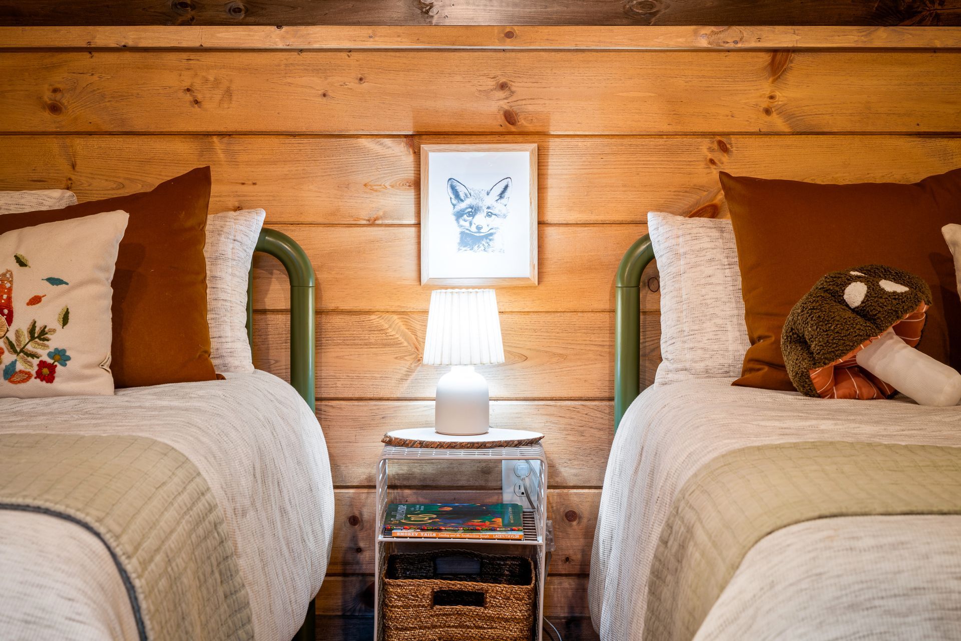 Two twin beds with wood paneling, a small lamp, and wall art in a rustic bedroom setting.