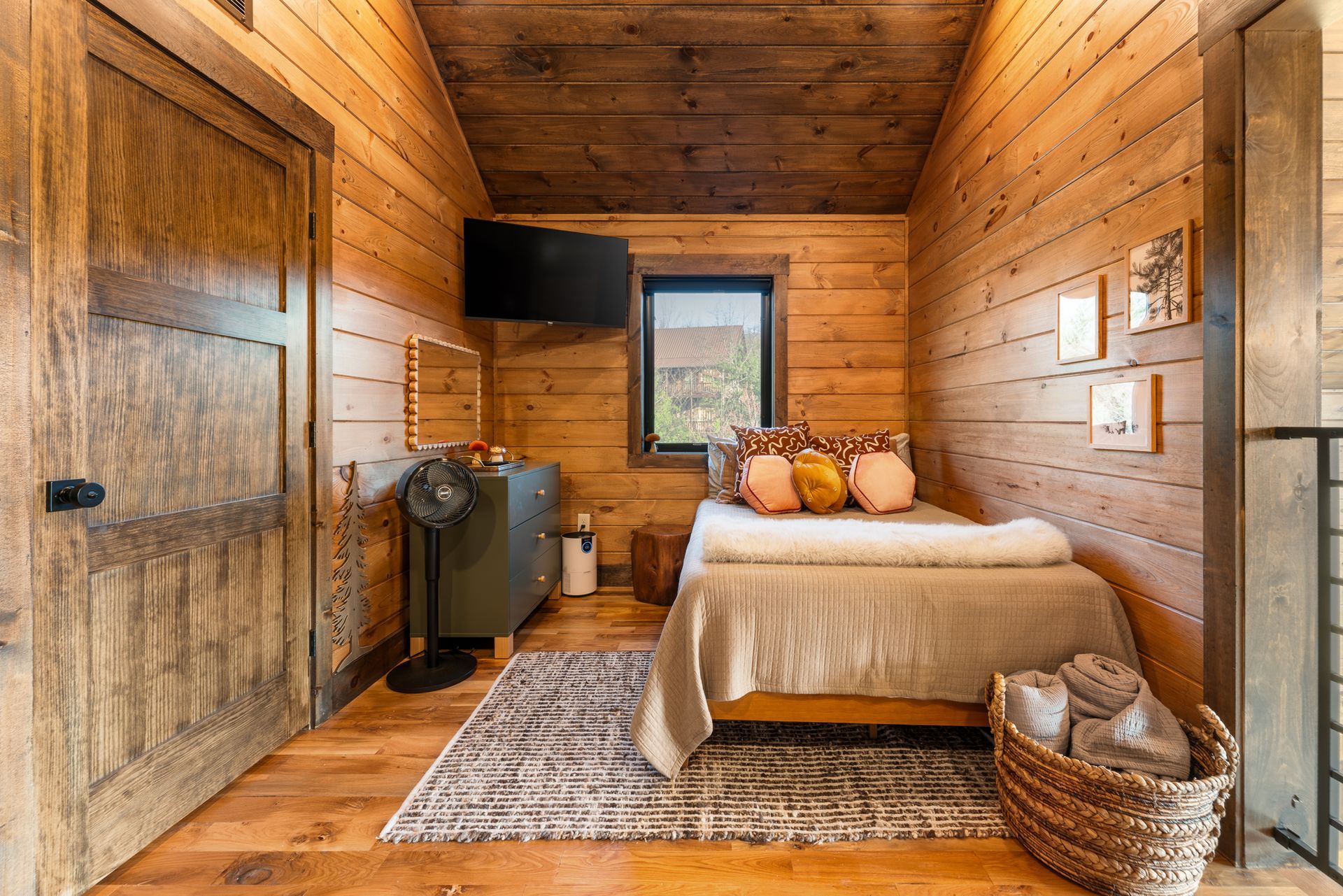 A rustic bedroom with wood-paneled walls, a bed, a small dresser with a TV above it, and a woven basket on a patterned rug.