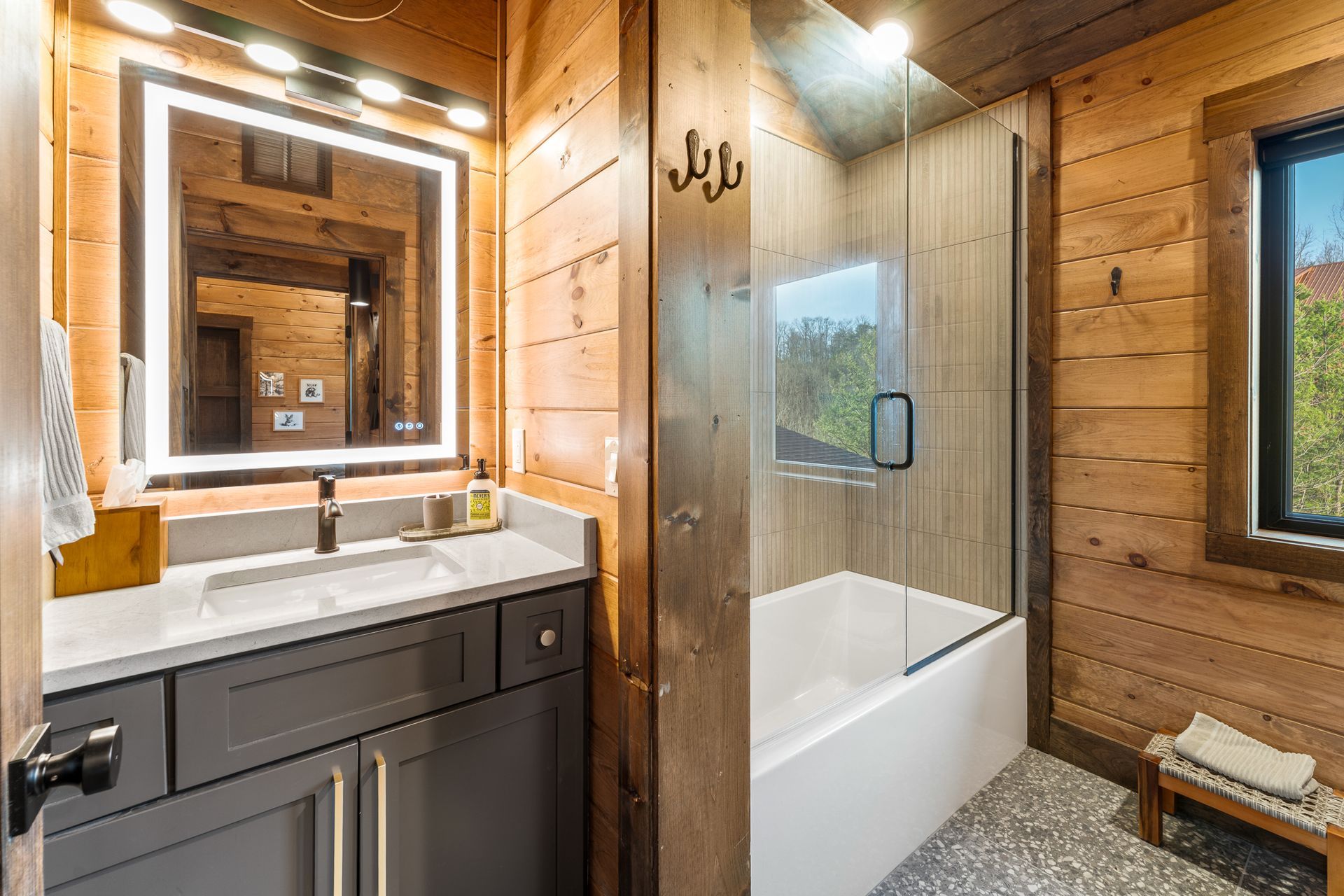 A rustic bathroom with wood walls, a dark grey vanity, a lighted mirror, and a walk-in shower with a glass panel.