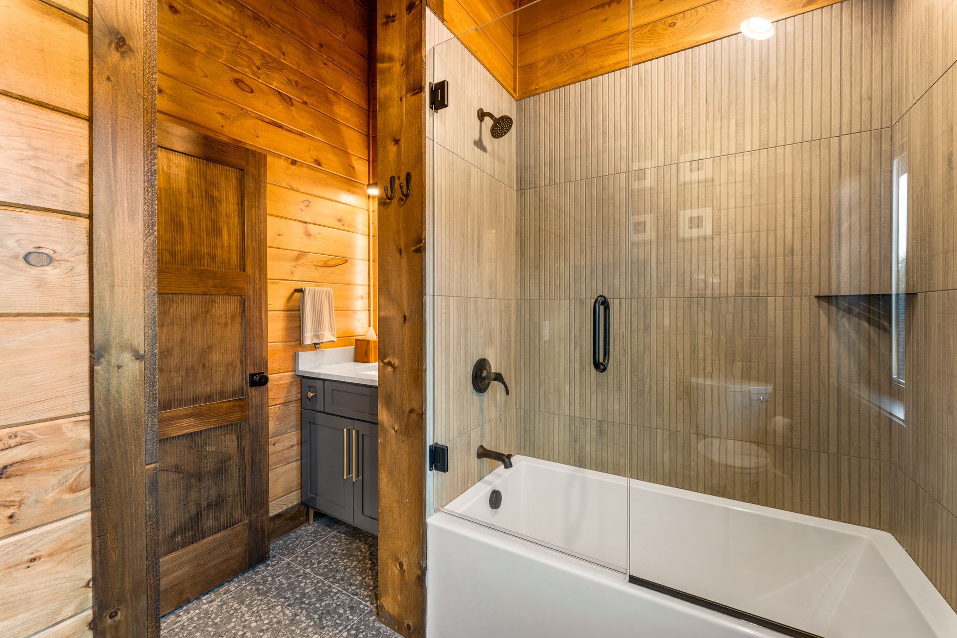 A rustic bathroom with wood walls, a grey cabinet, and a bathtub with a glass wall and shower head.