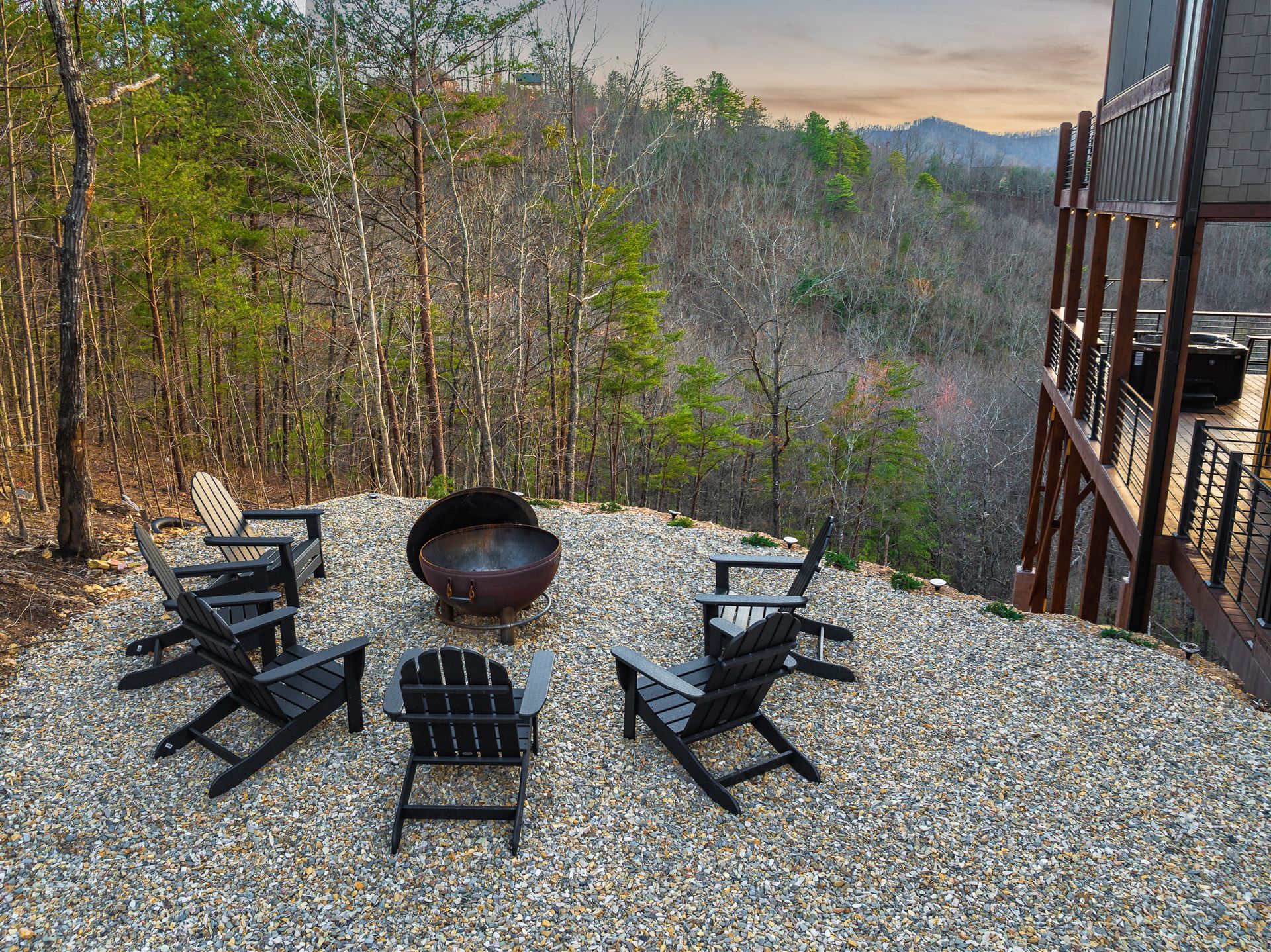 A fire pit with five black Adirondack chairs on a gravel patio overlooking a wooded mountain landscape at sunset.