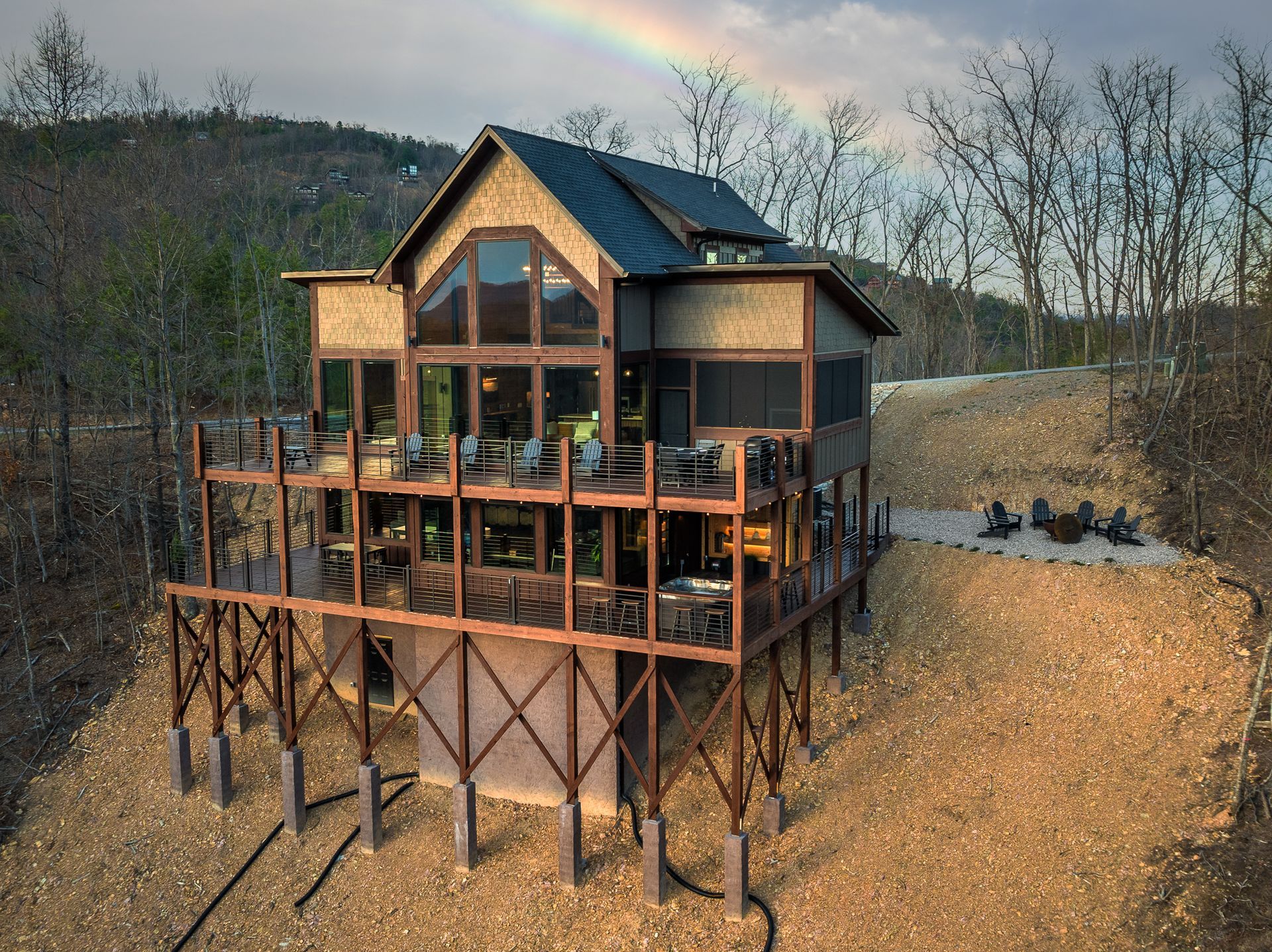 A multi-story wooden cabin perched on a hillside on stilts, featuring expansive windows and a deck under a faint rainbow.