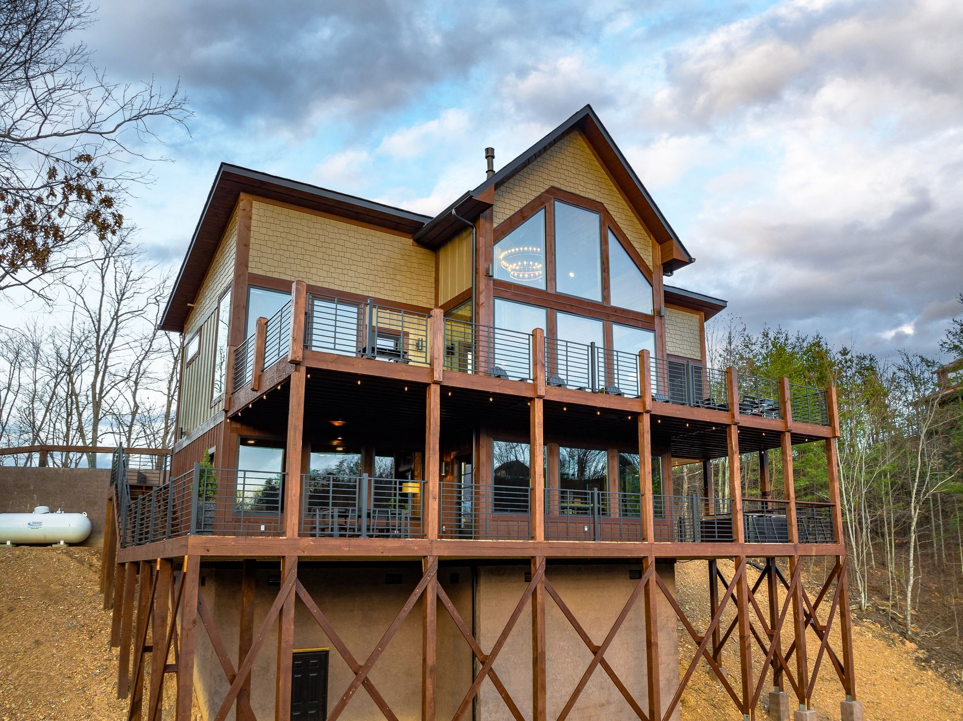A two-story cabin with a large glass facade and a wooden deck supported by stilts sits on a hillside against a cloudy sky.
