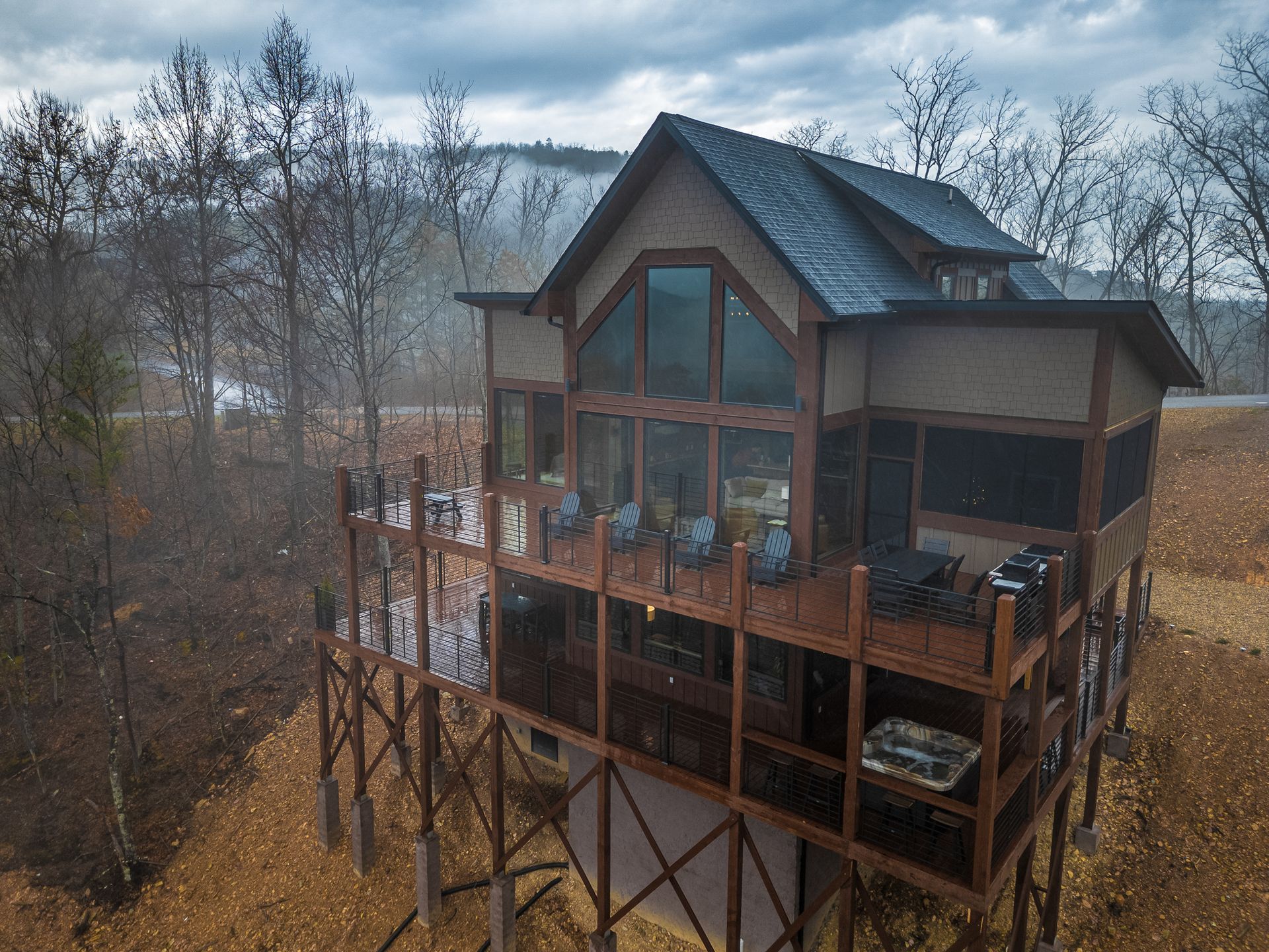 A wooden cabin with large windows and a tiered deck stands on a hillside surrounded by trees under a cloudy sky.