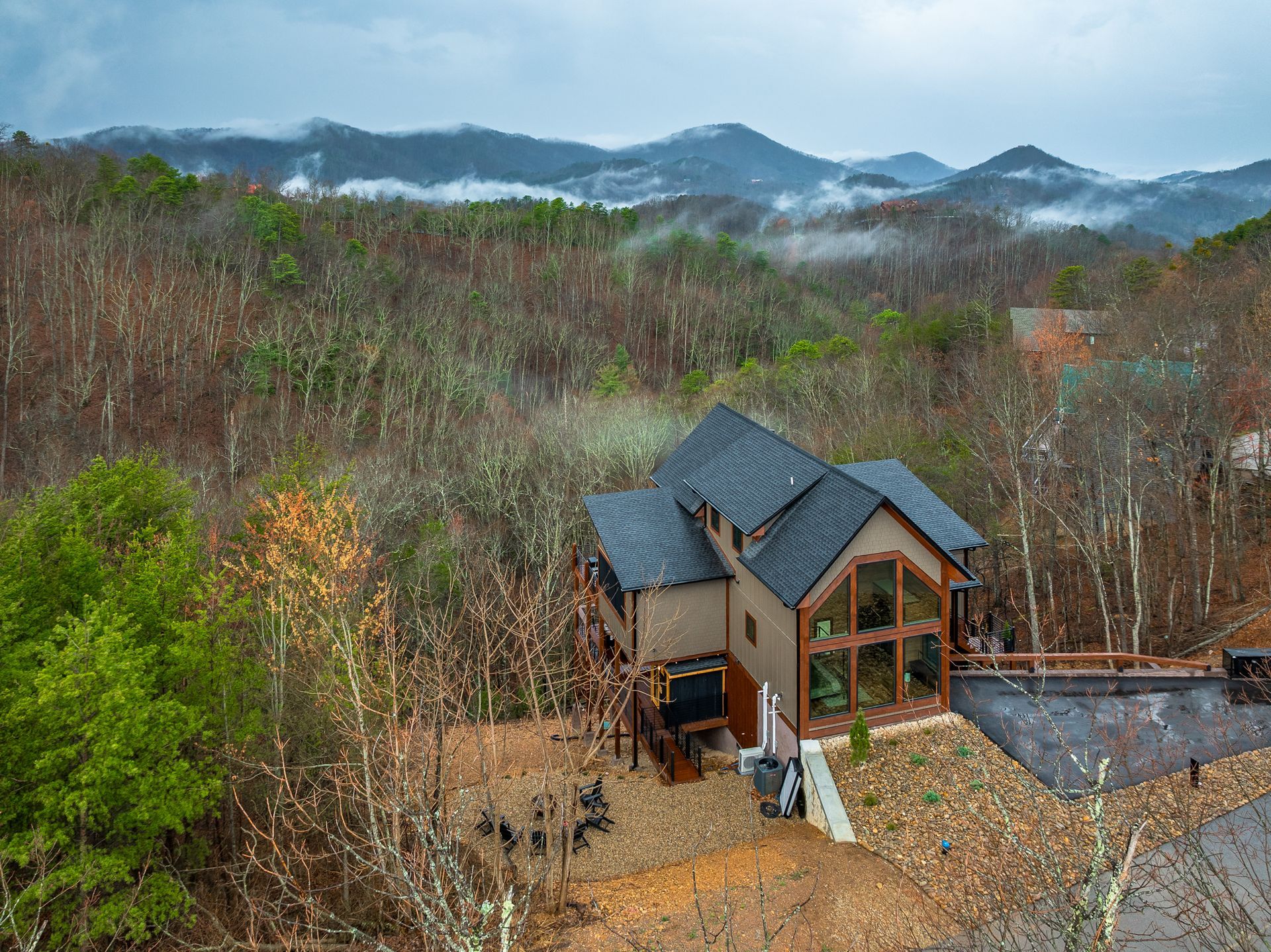 A multi-story wooden cabin with a dark roof sits nestled among trees, with misty mountains in the background.