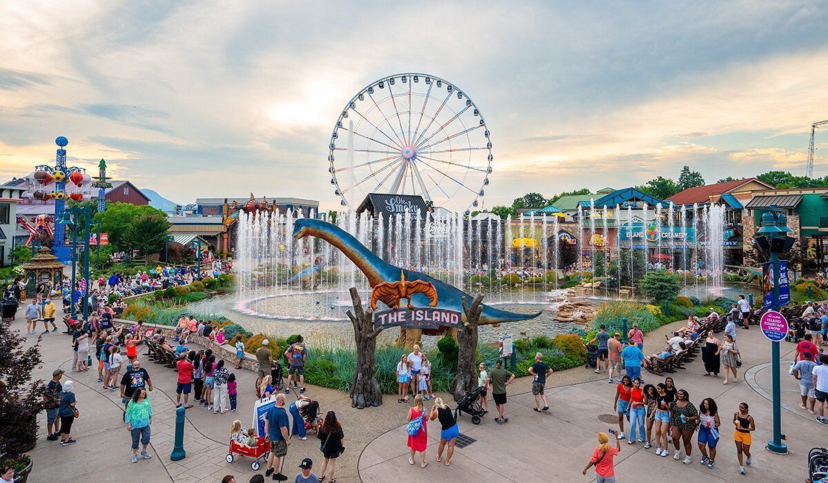 A large dinosaur statue sits in a fountain at an amusement park with a ferris wheel and crowds of people in the background.