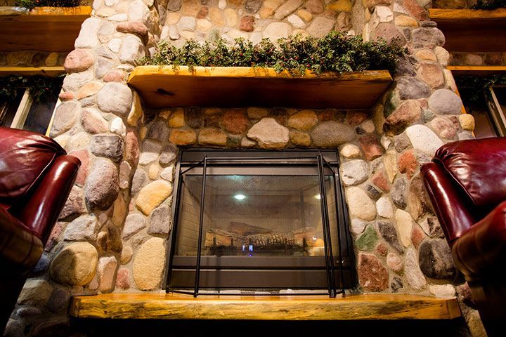 Stone fireplace with lit fire, wood mantel, and leafy greenery. Red leather chairs flank it.
