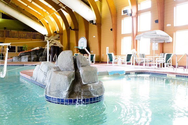 Indoor hotel pool with rock feature, arching ceiling, tables, chairs, and windows.