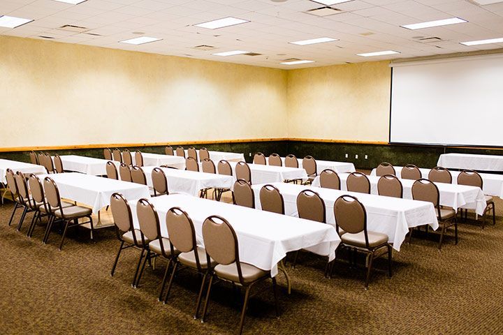 Conference room with rows of tables covered in white linens, brown chairs, and a projection screen.