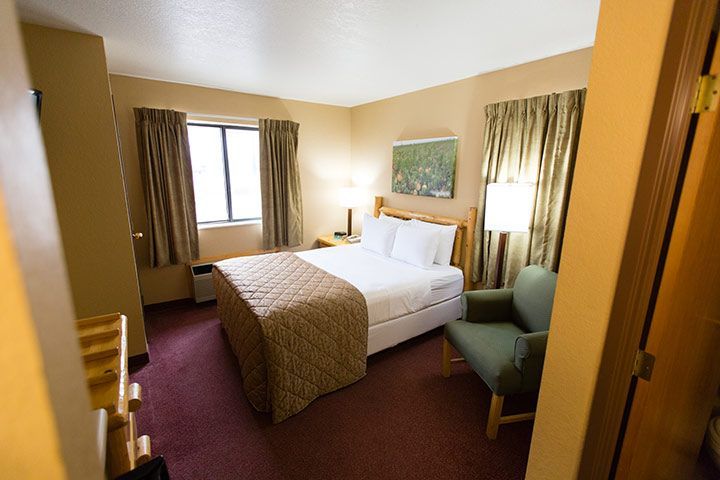 Hotel room with a bed, chair, and window. Beige walls, brown curtains, and a red carpet.