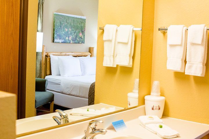 Bathroom mirror reflecting a hotel room with a bed, towels, and a sink. Yellow walls and white linens.