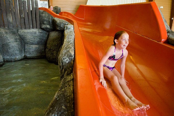Girl in a swimsuit slides down an orange water slide into a pool, smiling. Indoor water park setting.
