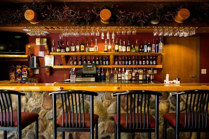 Bar with stone facade, wood countertop and shelves filled with bottles, stemmed glasses overhead, and black barstools.