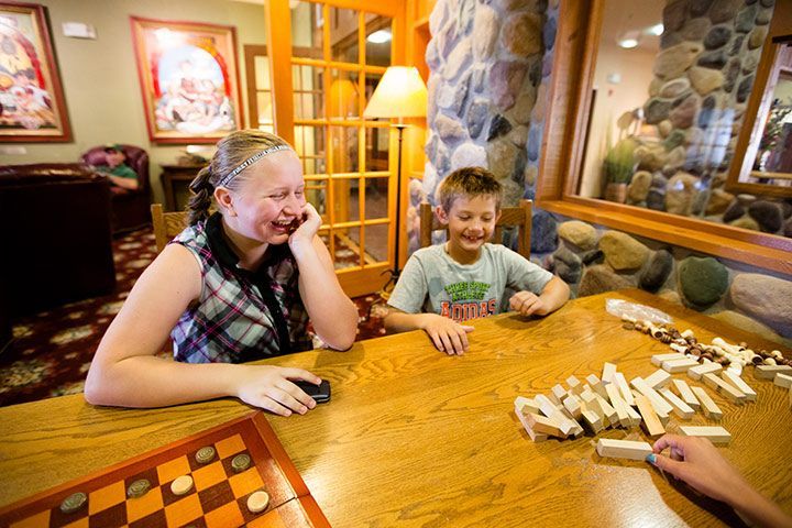 Two children playing a board game at a table. One laughs, looking at the other. Stone wall, warm lighting.