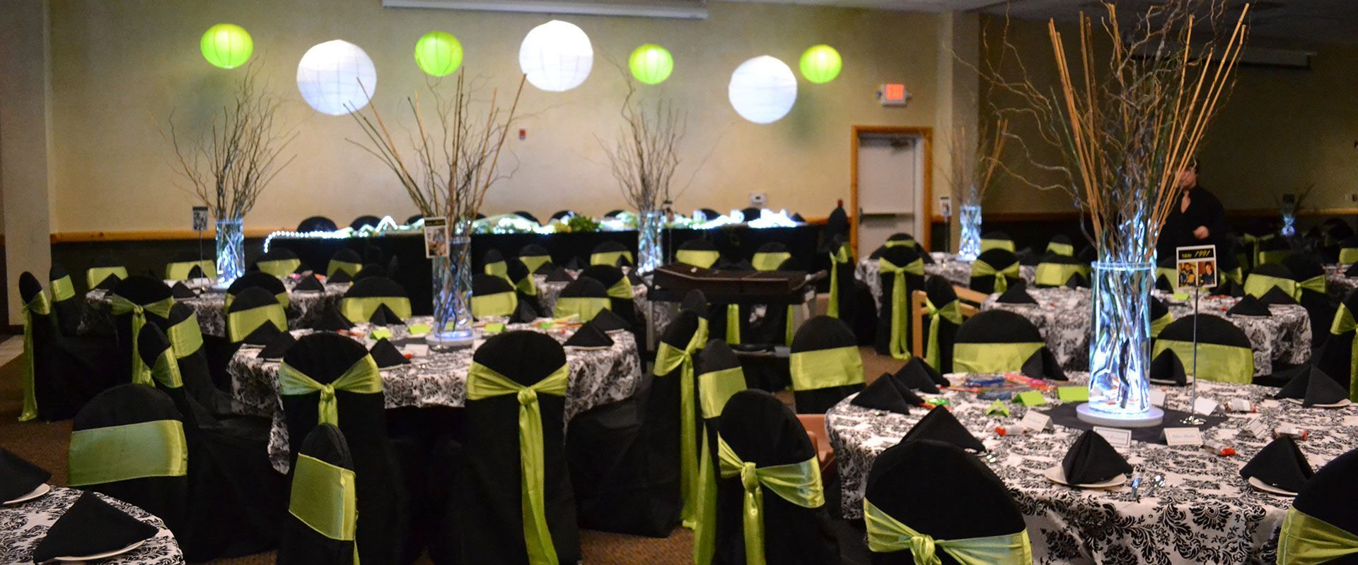 Reception hall decorated with black and white tablecloths, green chair sashes, and hanging paper lanterns.