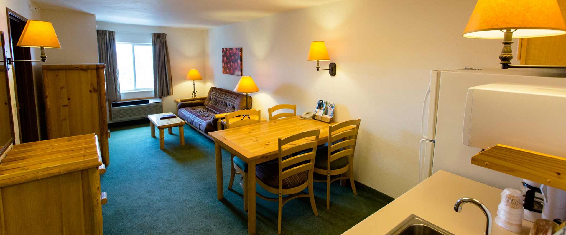 Cozy hotel suite with seating area, dining table, kitchen counter, and multiple warm-toned lamps.