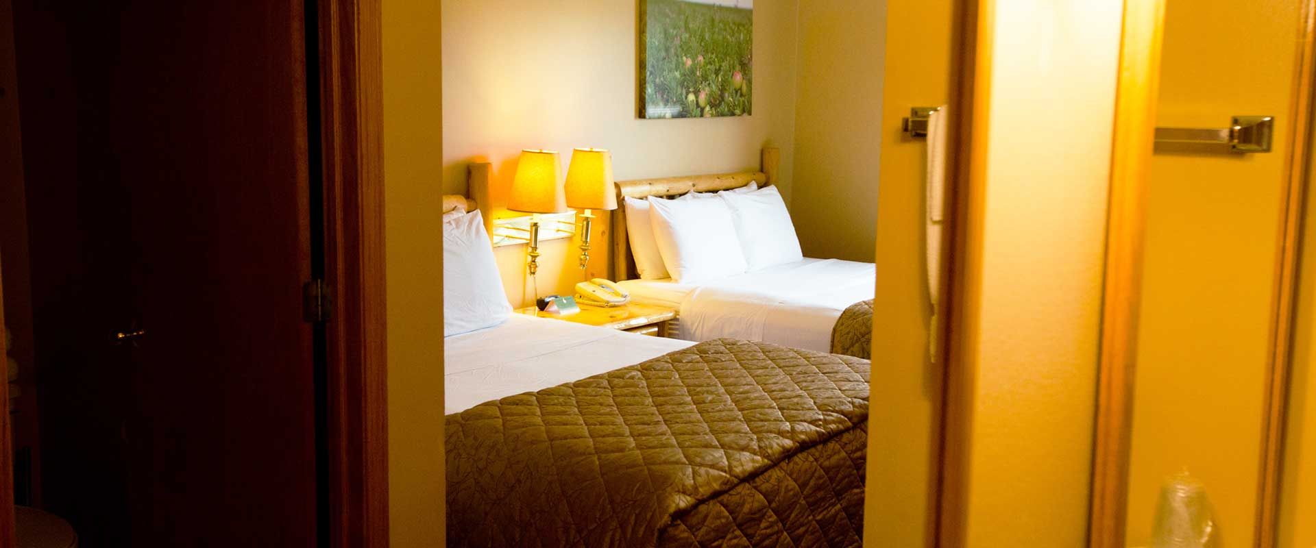 View of a hotel room with two beds through a doorway. Yellow walls, lamps, and art on the wall.